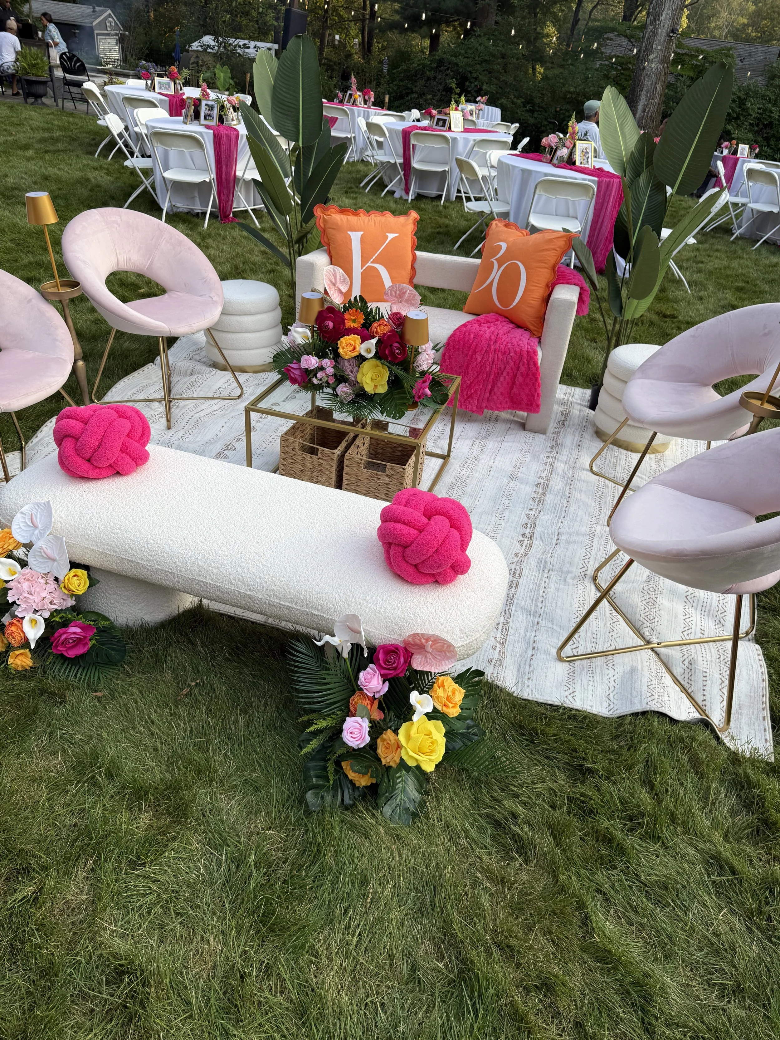 Outdoor event setup with pink, white, and orange decorations. Pink velvet chairs, cream sofa with decorative pillows marked with 'K' and 'O', surrounded by colorful floral arrangements. White round tables with pink tablecloths and floral centerpieces are in the background, set on a grassy lawn with trees.