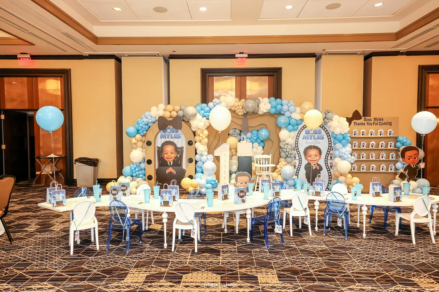 Blue-themed birthday party setup with balloon arch, large photos of a young boy, birthday banners, tables with party favors, and character cutouts.