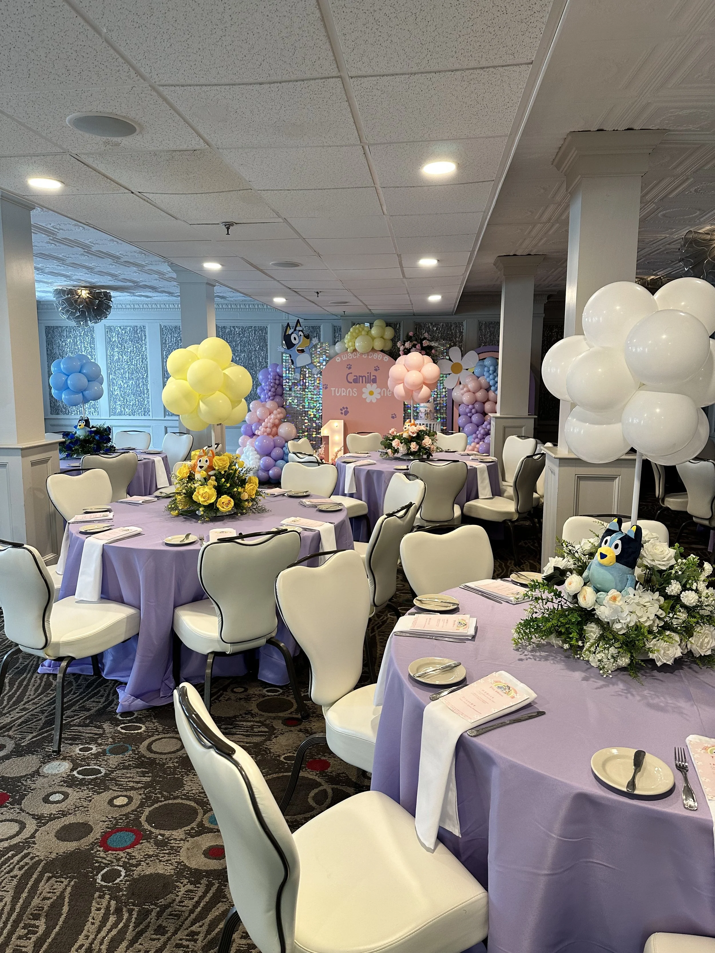 Decorated banquet hall for a child's birthday party with purple tablecloths, balloon arrangements, and a backdrop featuring a cartoon character and a sign that reads "Camila turns one".