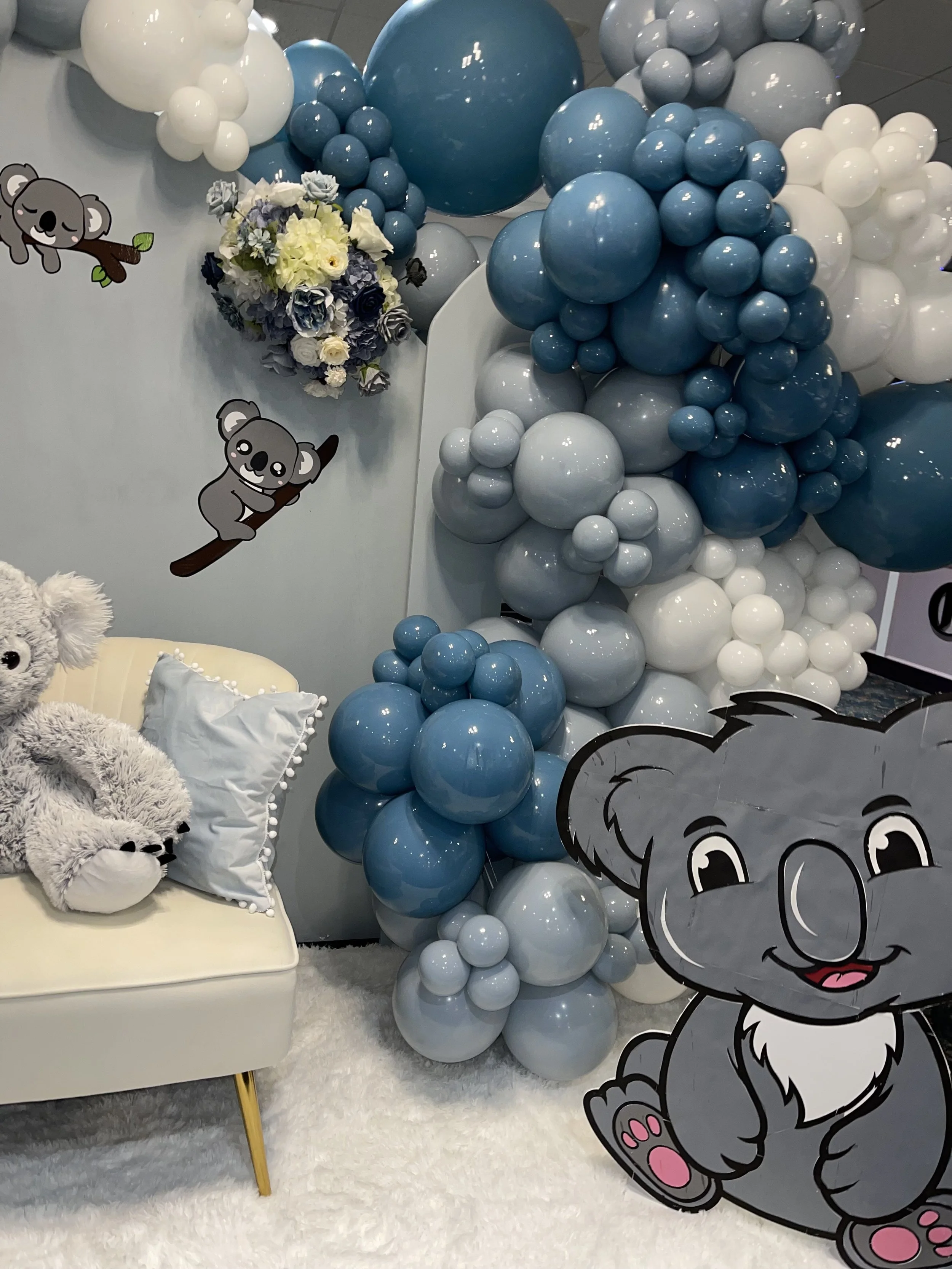 Baby shower decoration with koala theme, featuring balloons in shades of blue, white, and gray, a plush koala bear on a white chair, and cartoon koala cutouts on the wall.