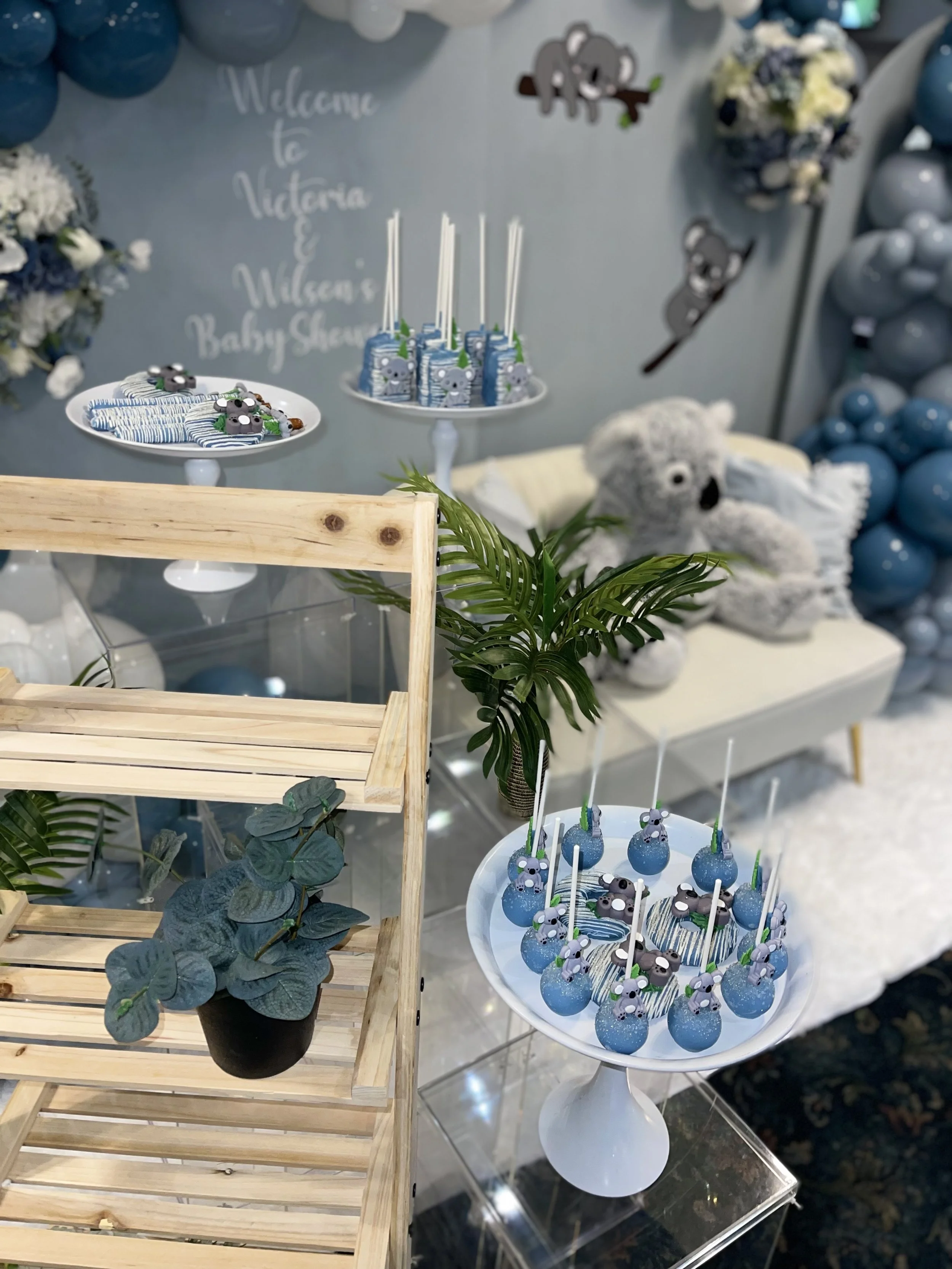 Baby shower setup with koala-themed decorations, including treats and cake pops featuring koalas, and a large teddy bear, with a backdrop that says "Welcome to Victoria & William's Baby Shower".