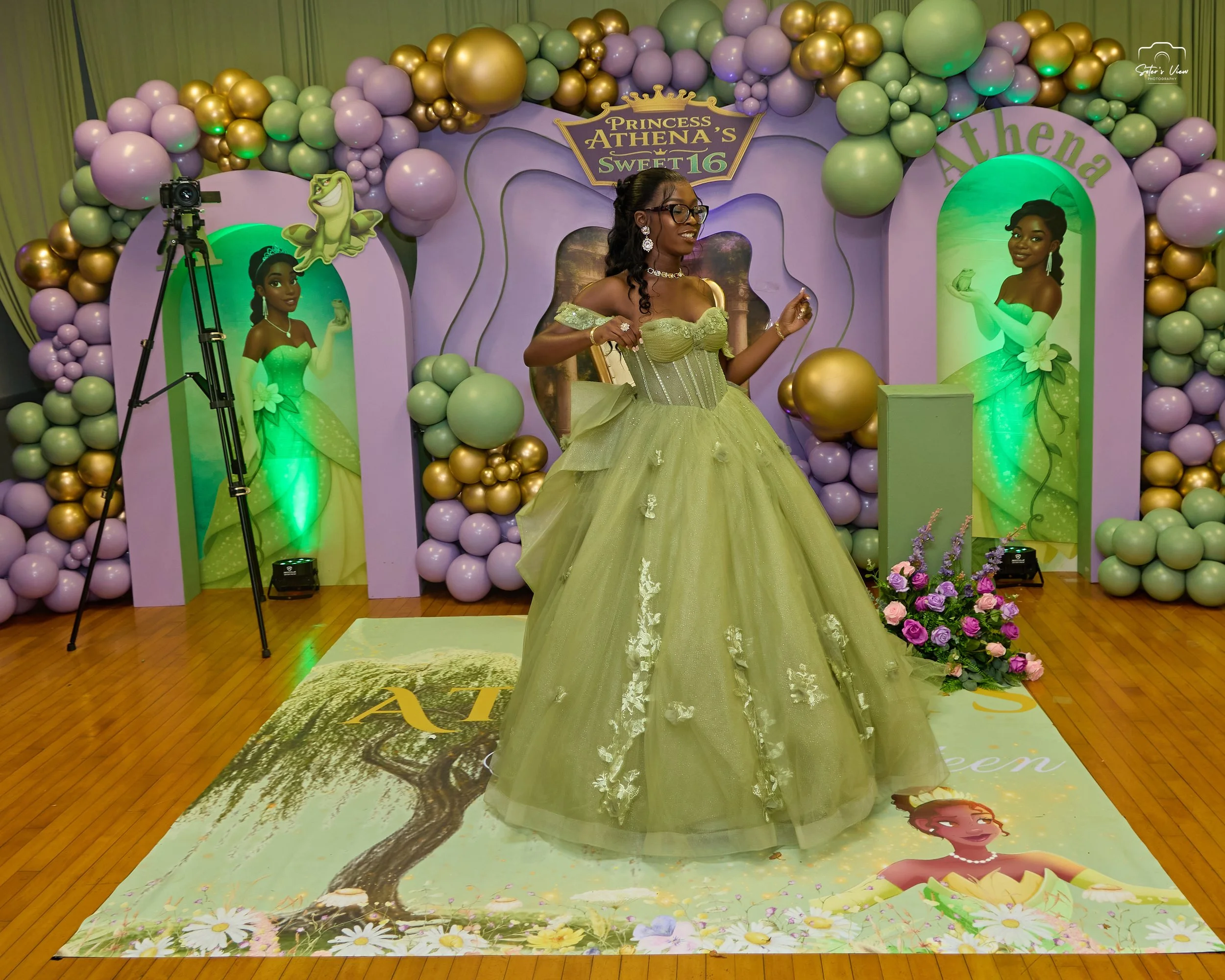A girl dressed in a green princess gown with floral details, standing on a themed floor mat featuring a princess and a tree, at her birthday party celebration. The background includes pastel-colored balloon decorations, animated princess images, and a banner reading 'Princess Athena's Sweet 16'.