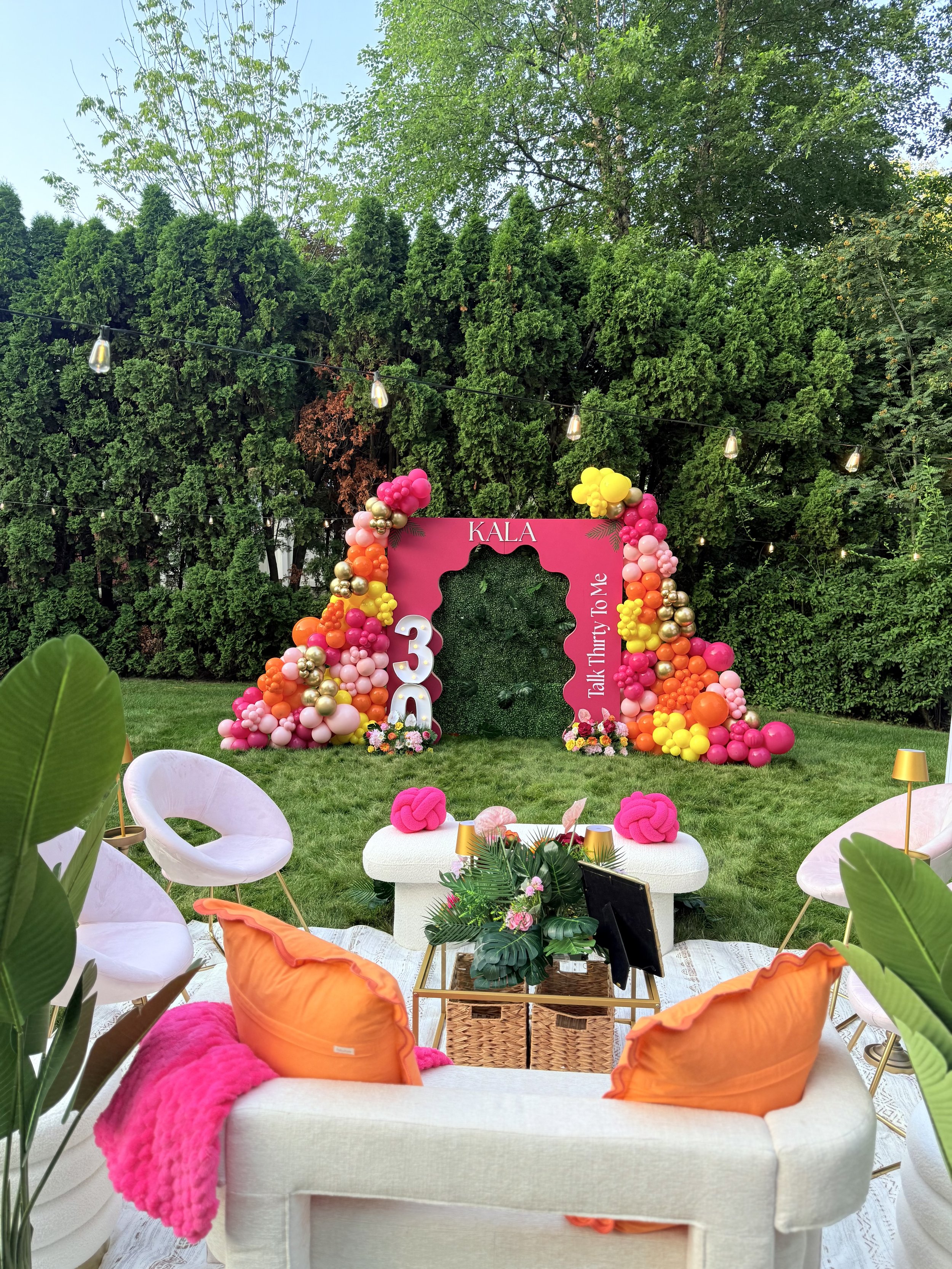 Outdoor birthday party setup with a decorated backdrop featuring pink, yellow, orange, and gold balloons, and a green hedge wall in the background. There are white lounge chairs with orange pillows, a white cushioned bench with pink decorations, and a small table with a floral arrangement and lanterns. String lights are hanging overhead.