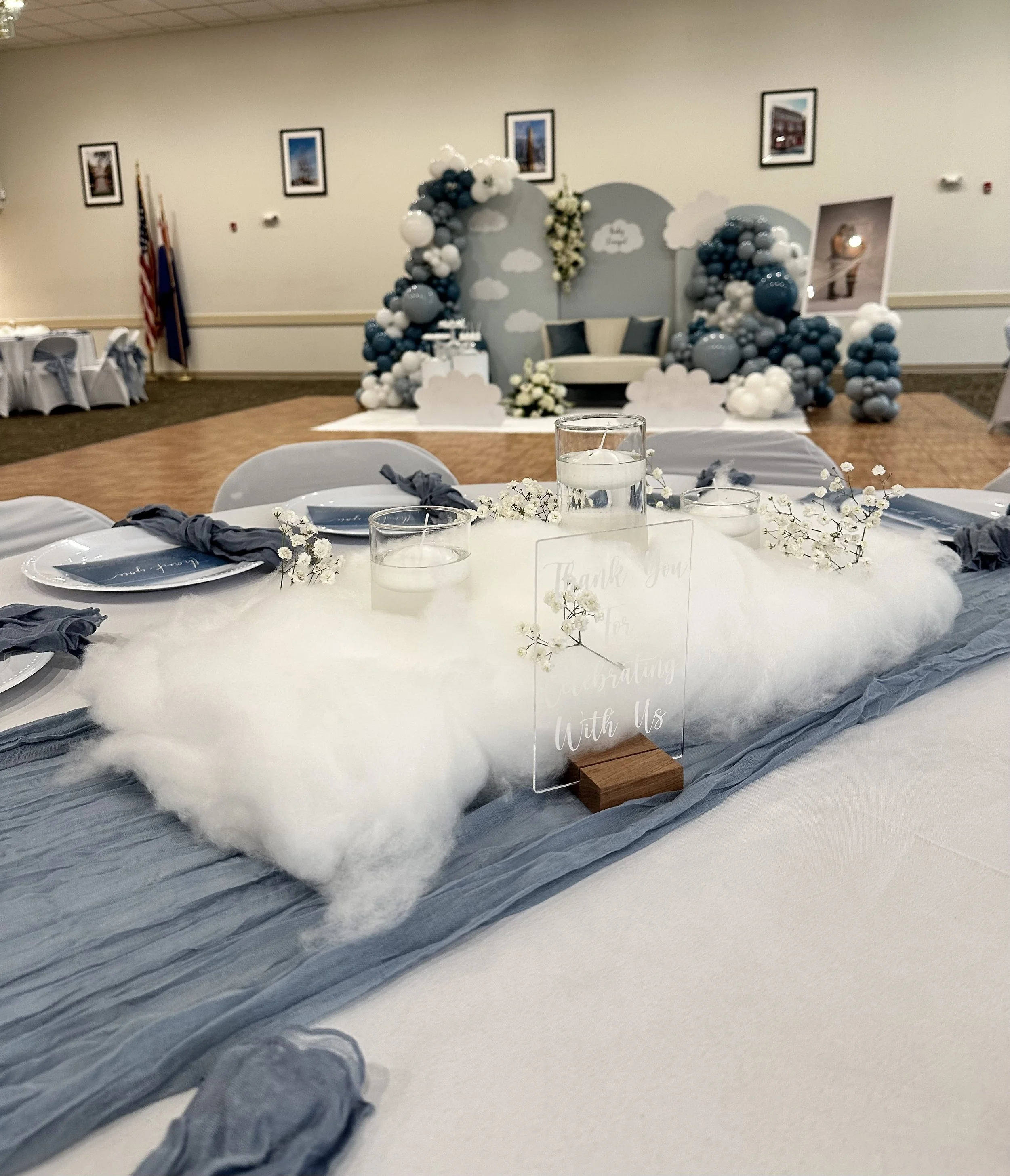 Wedding reception table with navy blue and white decorations, flowers, candles, and a thank you sign in foreground; a decorated stage with balloons, clouds, and seating area behind.