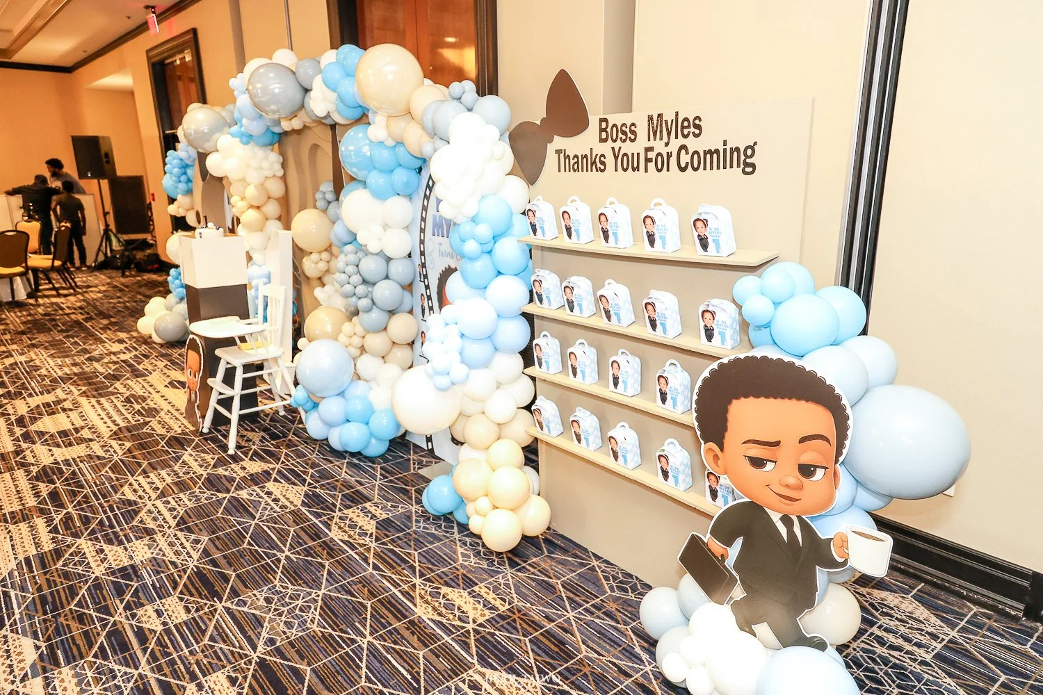 Decorative event display with blue, white, and beige balloons, a cardboard character holding a coffee mug and a briefcase, and a sign that reads "Boss Myles Thanks You For Coming." The display includes a shelf with favor boxes featuring a cartoon image of a person.