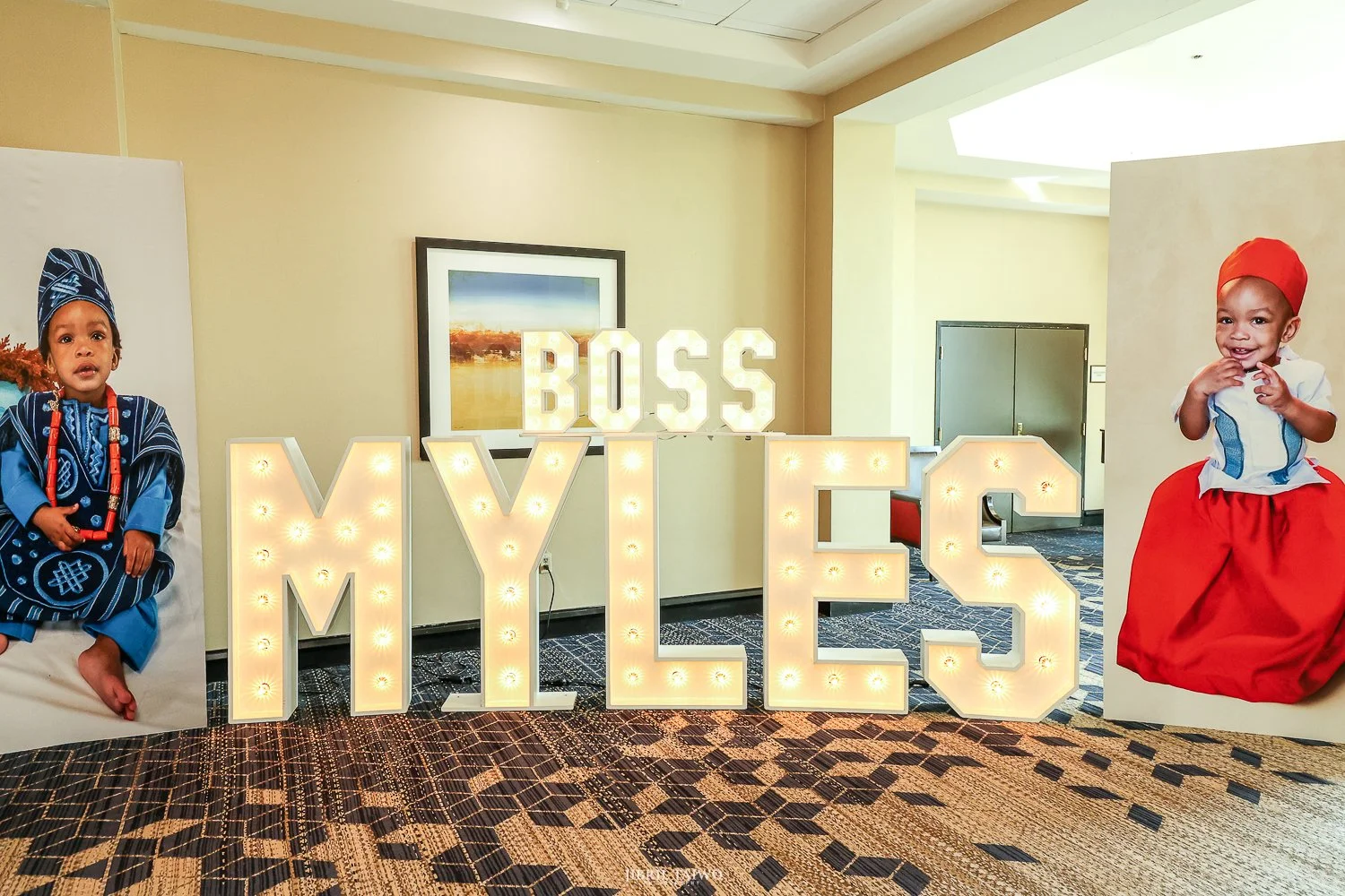 Two children in traditional African attire, one on each side of large illuminated letters spelling 'MY ELE' with 'BOSS' on top, inside a hotel or event venue. The child on the left is wearing blue patterned clothing, the child on the right is wearing a white shirt and a red skirt with a red headwrap.