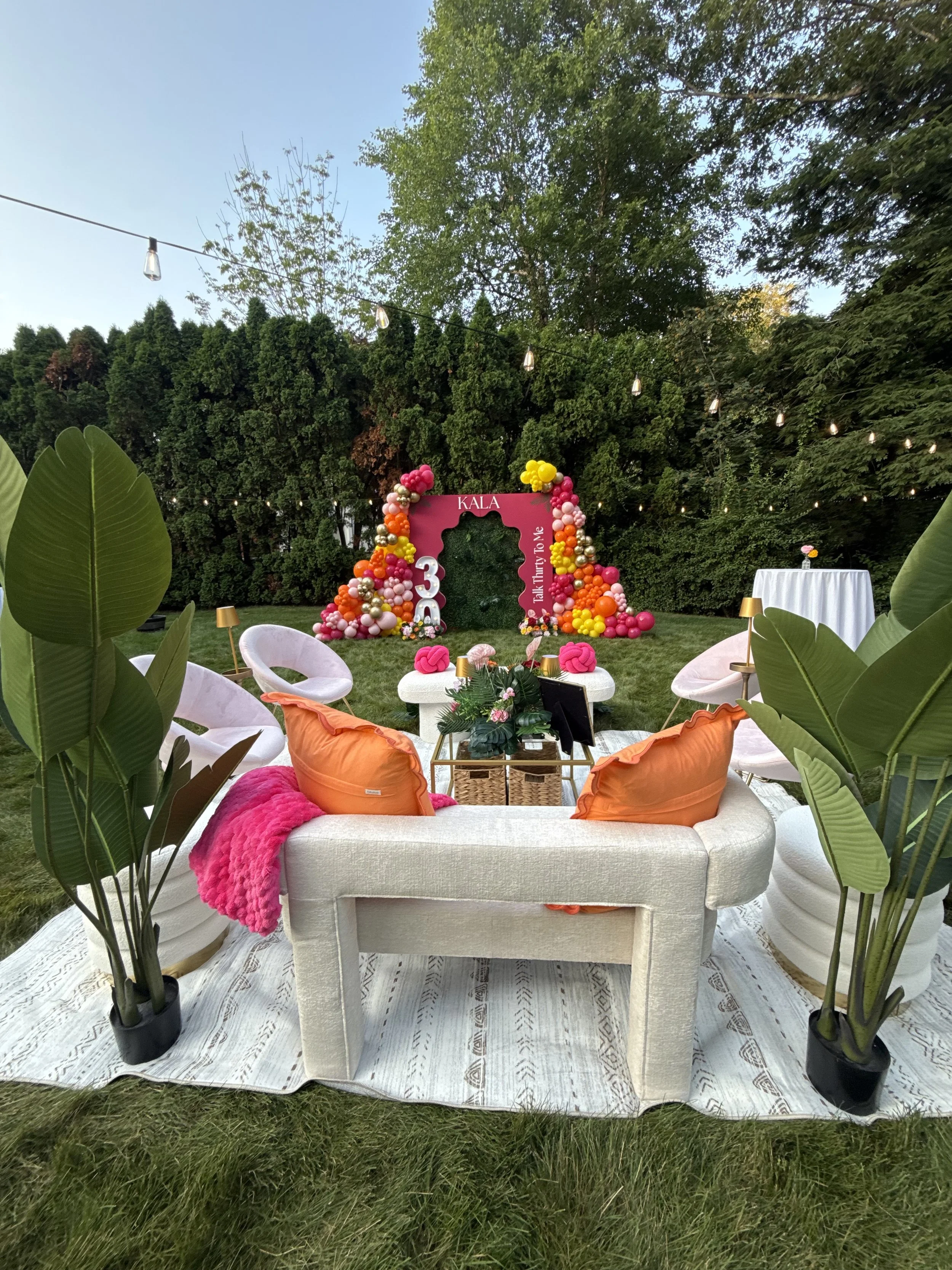 Outdoor birthday party setup with pastel furniture, pink and orange balloons, large potted plants, a pink backdrop with balloons, and a grassy yard with trees.