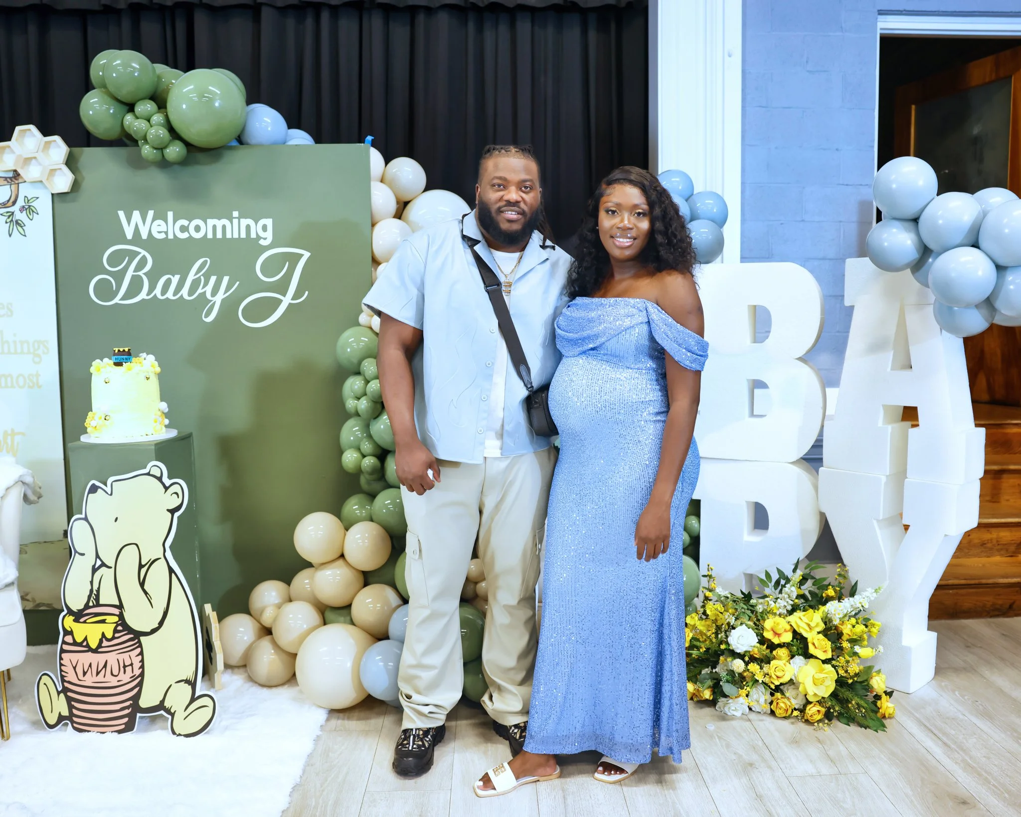 A couple stands together at a baby shower next to a green sign that says 'Welcoming Baby J' with balloons and decorations in the background. The woman is wearing a blue dress and the man is dressed casually.