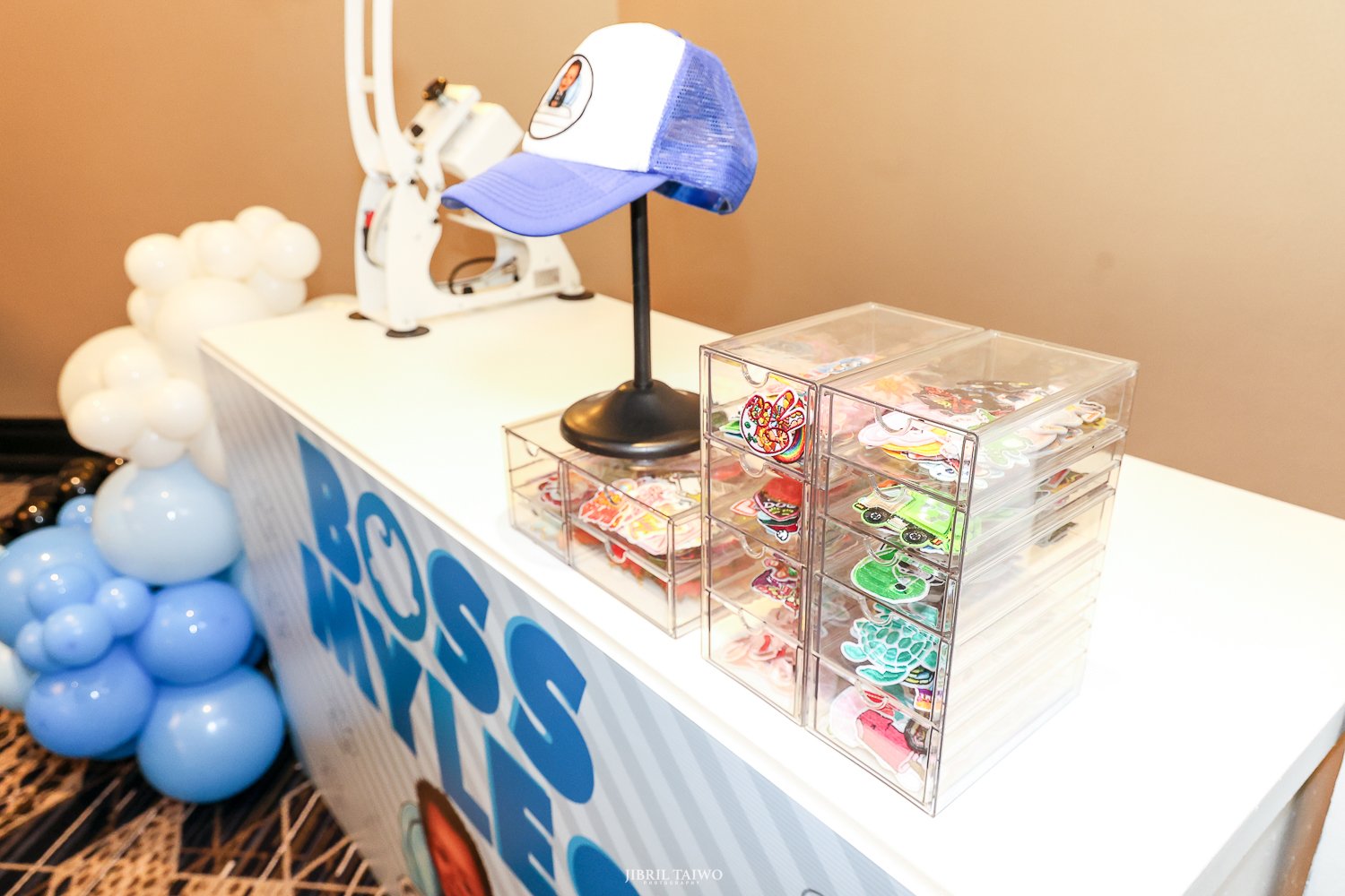 A table display with a blue and white baseball cap, clear plastic containers filled with colorful stickers, and a small stand with a baseball cap on top. Decorations include clusters of blue and white balloons on the side of the table.
