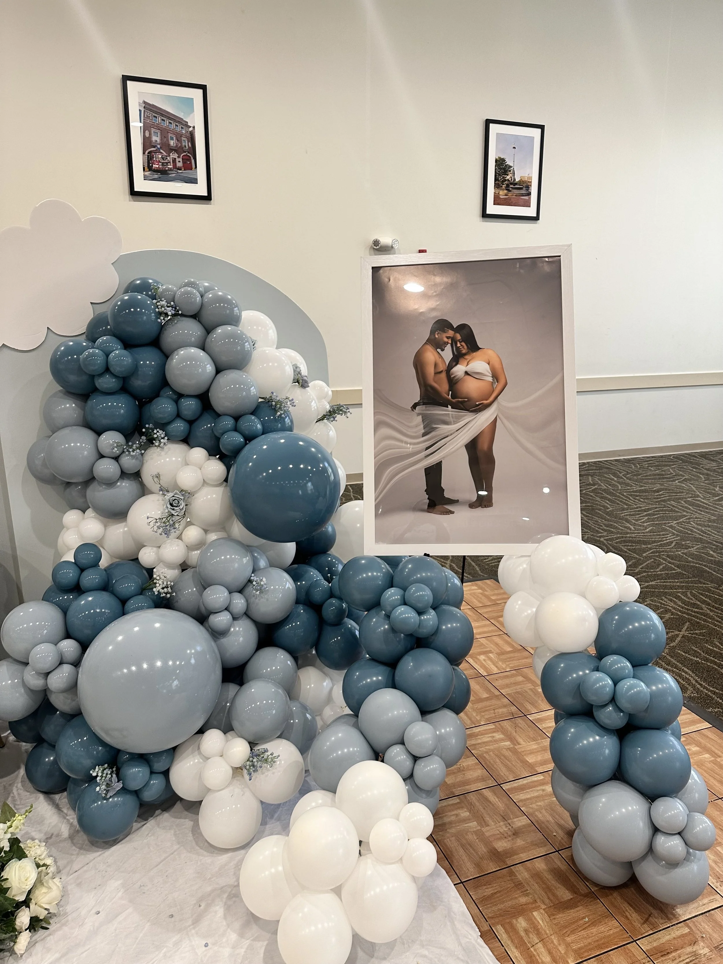 Decorative display with a cluster of blue, white, and gray balloons next to a framed pregnancy portrait of a pregnant woman and a man, with two small framed photographs hanging on the wall in the background.