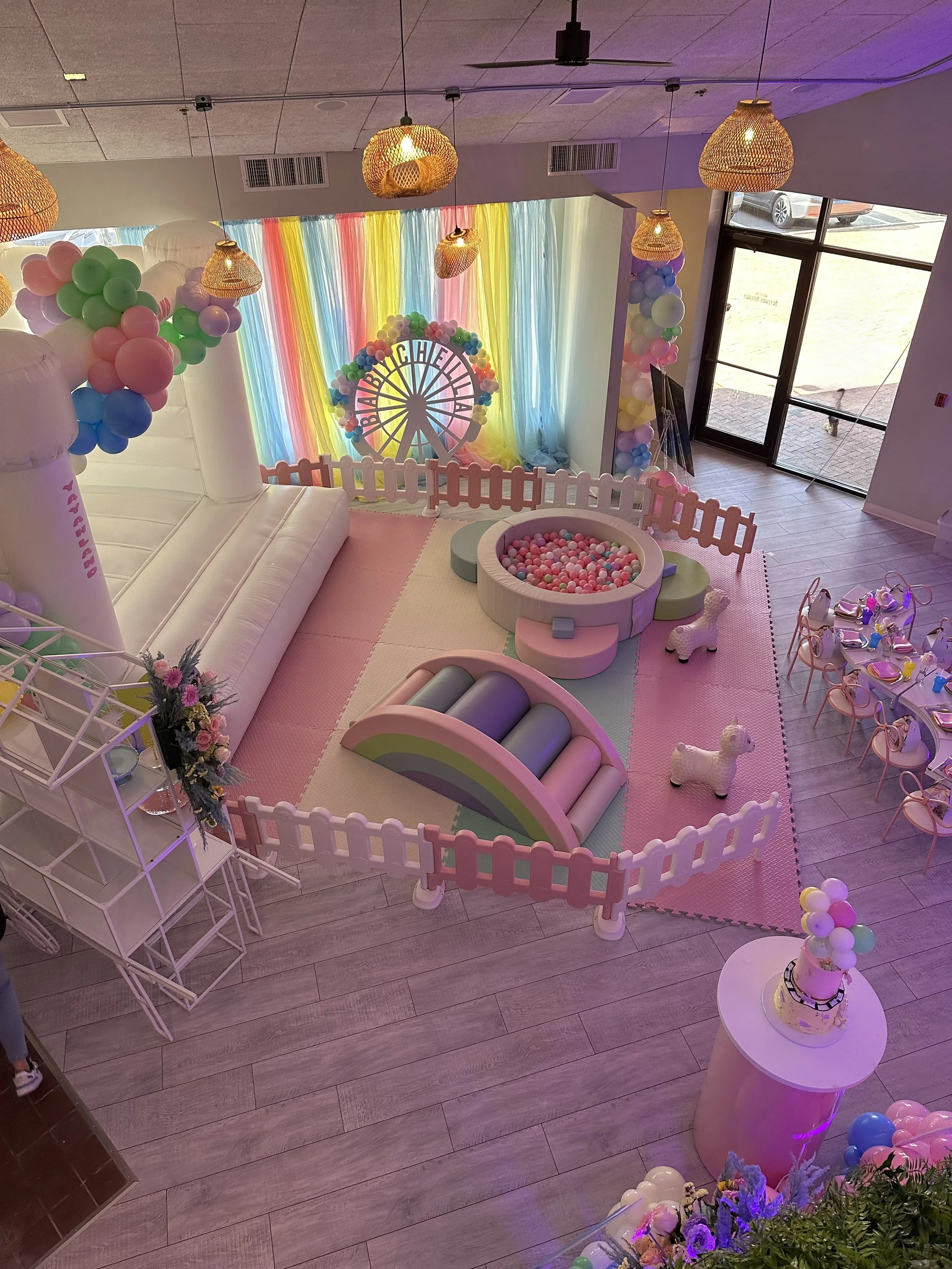 Colorful indoor party space decorated with balloons, a rainbow-colored backdrop, a small fenced play area with a ball pit, and tables for children with chairs, set up for a celebration or birthday party.