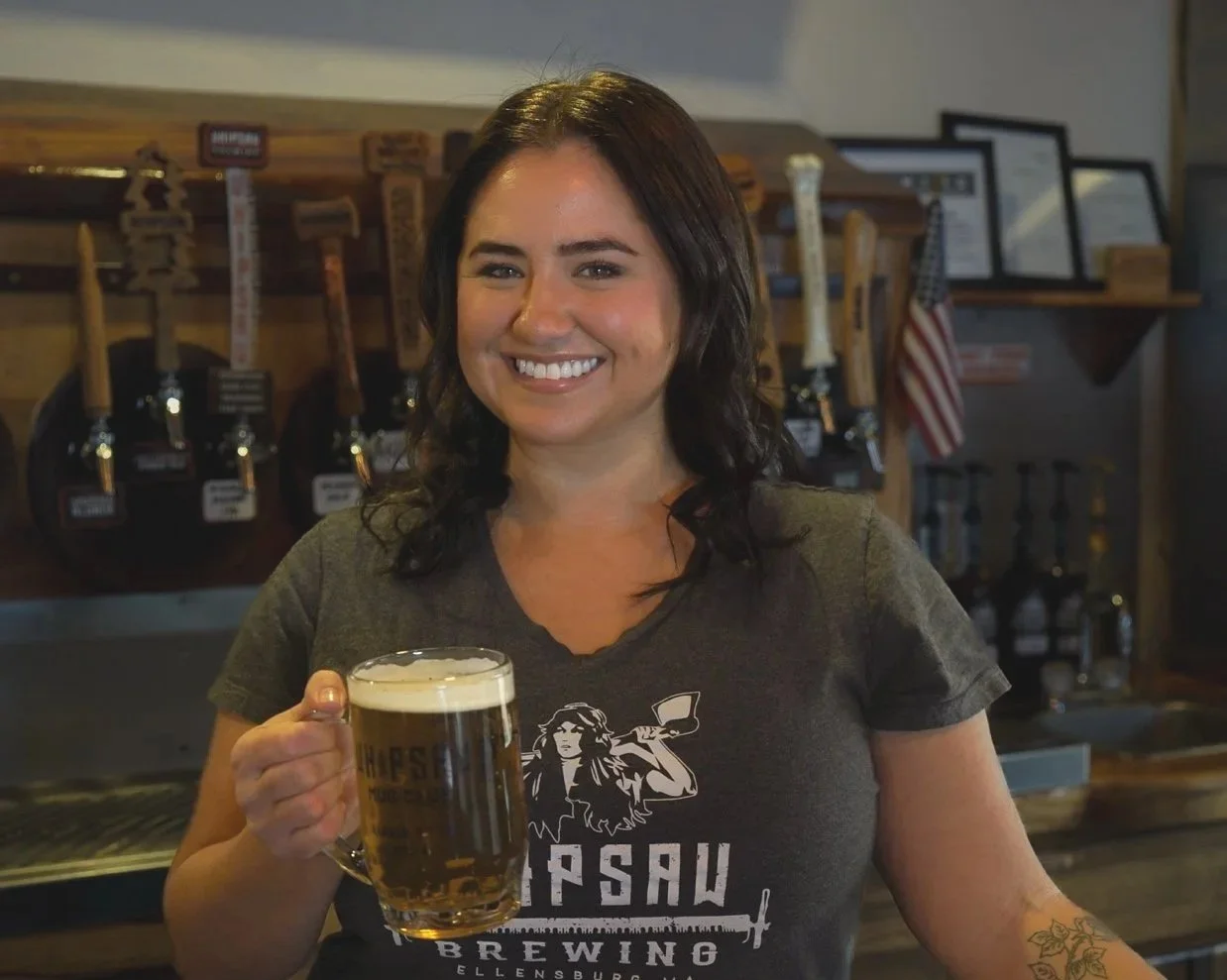 A photo of Nikki, Tap Room Server at Whipsaw Brewing in Ellensburg, Washington, smiling with a pounder of locally brewed Whipsaw Stumpblower beer.