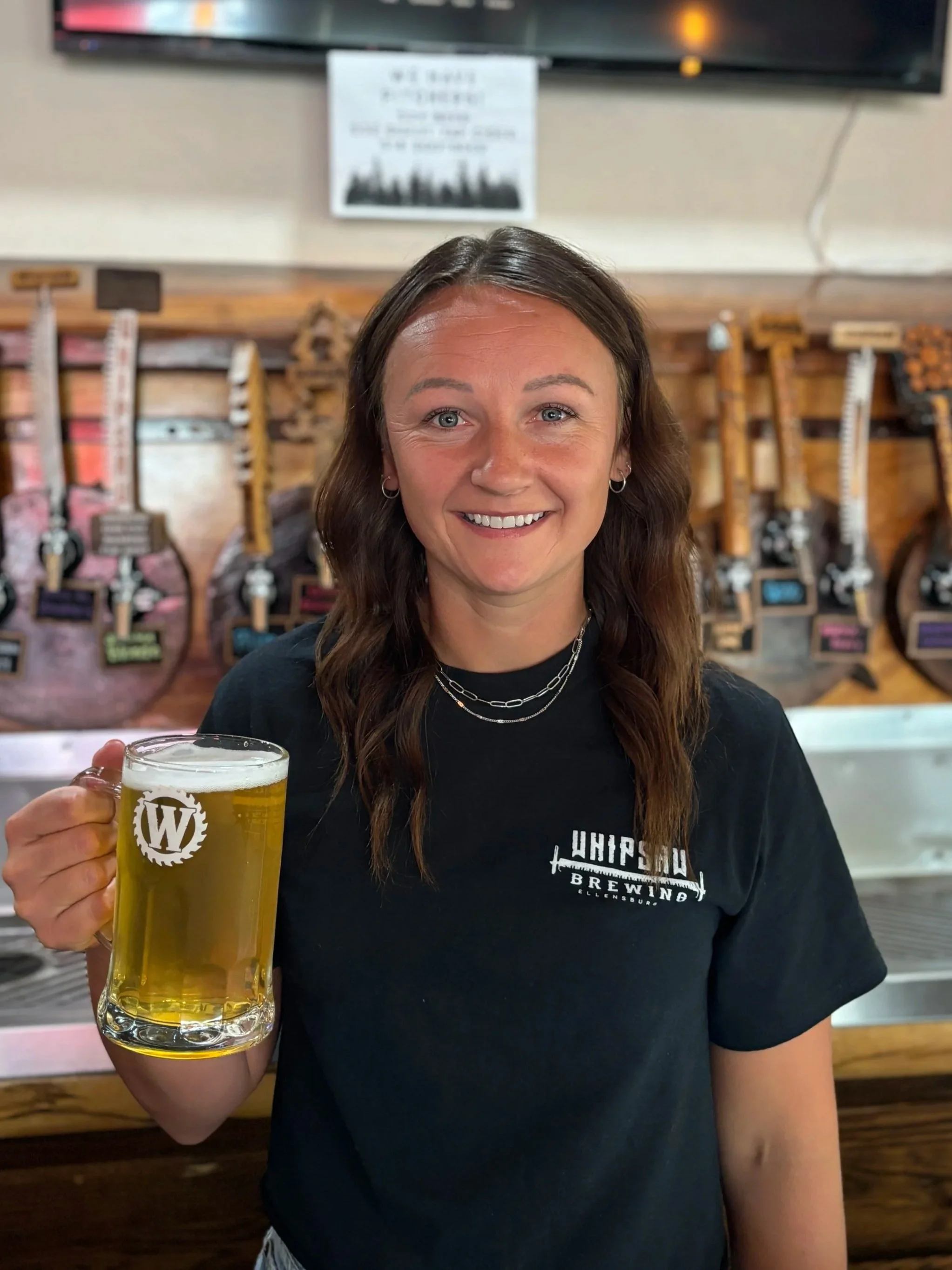 A photo of Bailie, Tap Room Server at Whipsaw Brewing in Ellensburg, Washington, smiling with a mug of locally brewed Whipsaw Smashed Blonde beer.