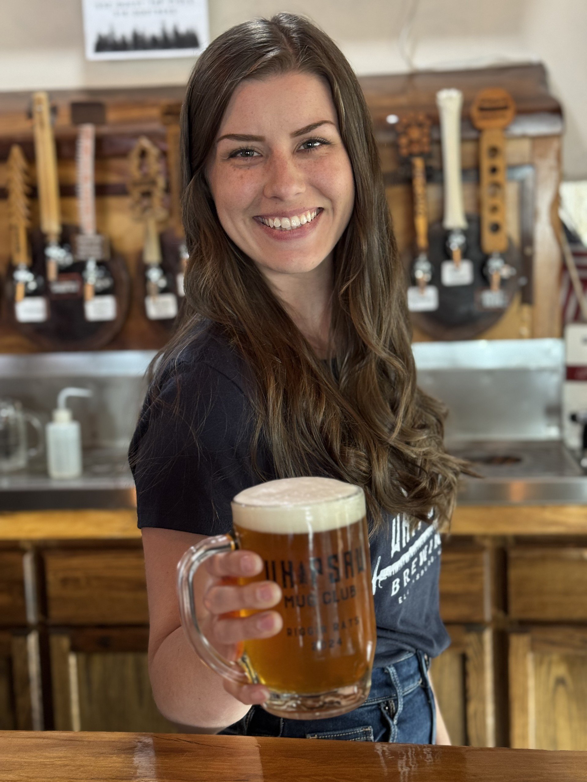 A photo of Jaelin, Tap Room Lead at Whipsaw Brewing in Ellensburg, Washington, smiling with a mug of locally brewed Whipsaw Smashed Blonde Stumpblower beer.