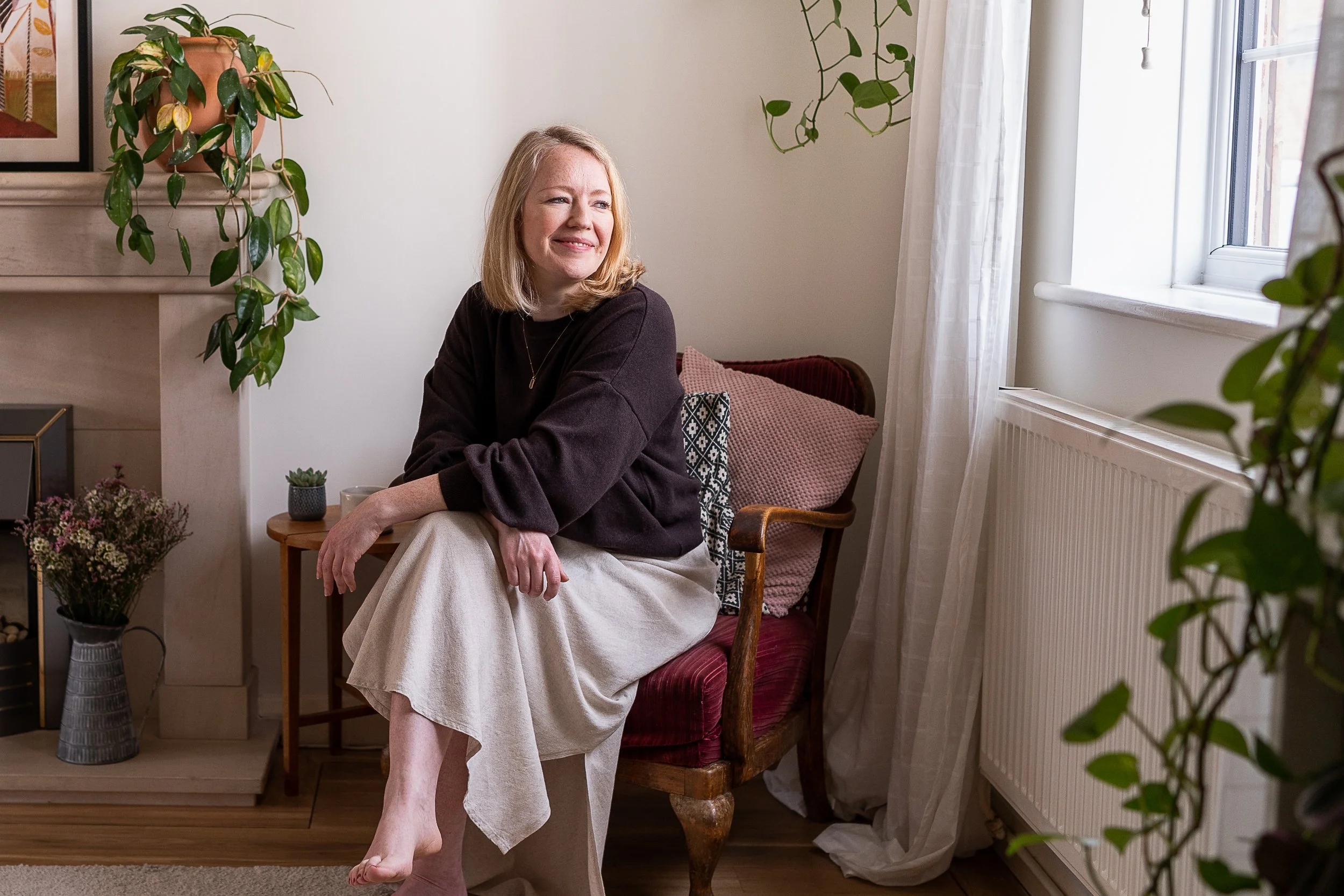 Lauren Johns, The Should I Stay Or Should I Go Coach, smiling with blonde hair sitting by a window in a cosy, flower-filled room, wearing a black top and a cream skirt.