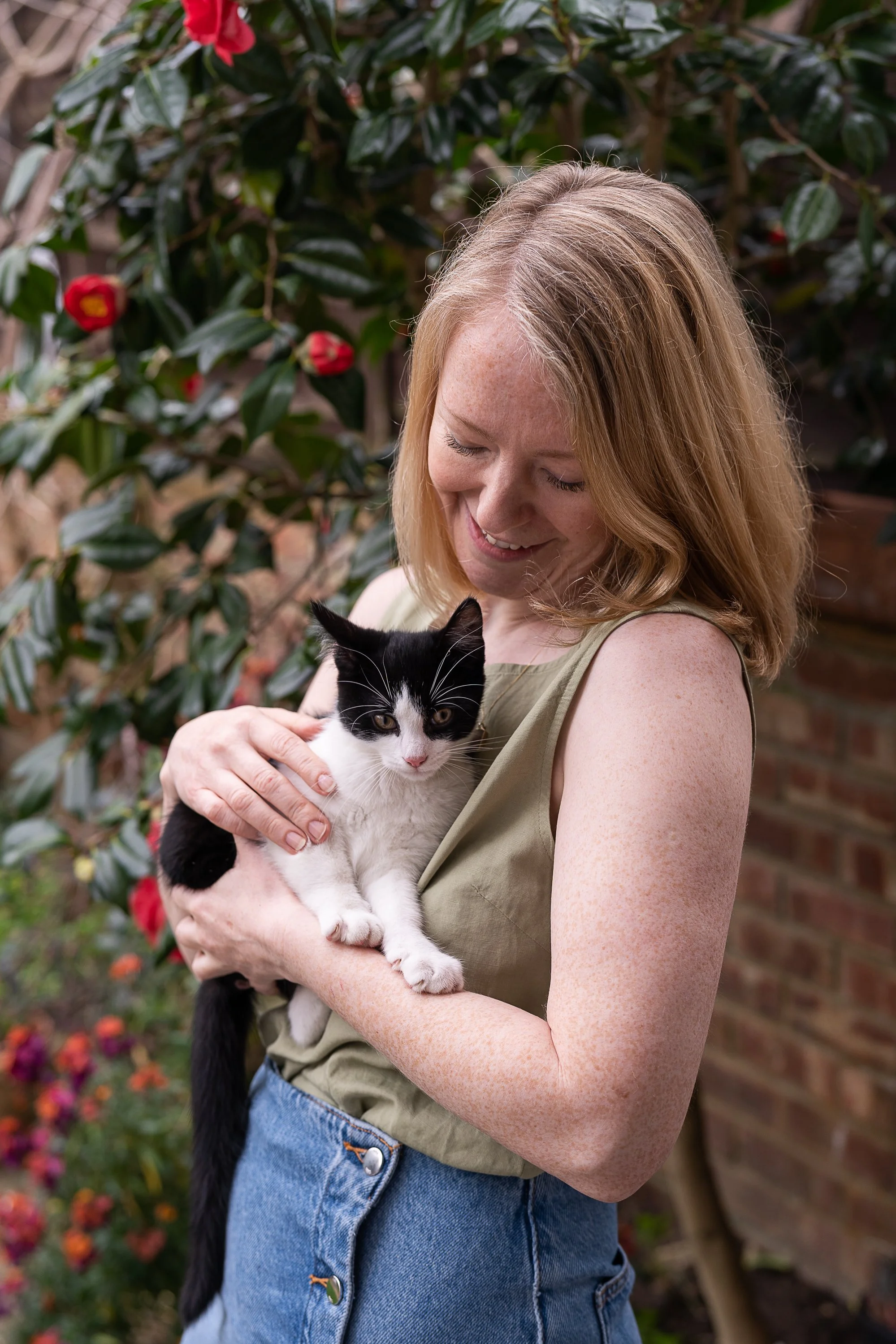 A woman with red hair, wearing a sleeveless top and jeans, holding a black and white cat outdoors near a brick wall and a flowering shrub.
