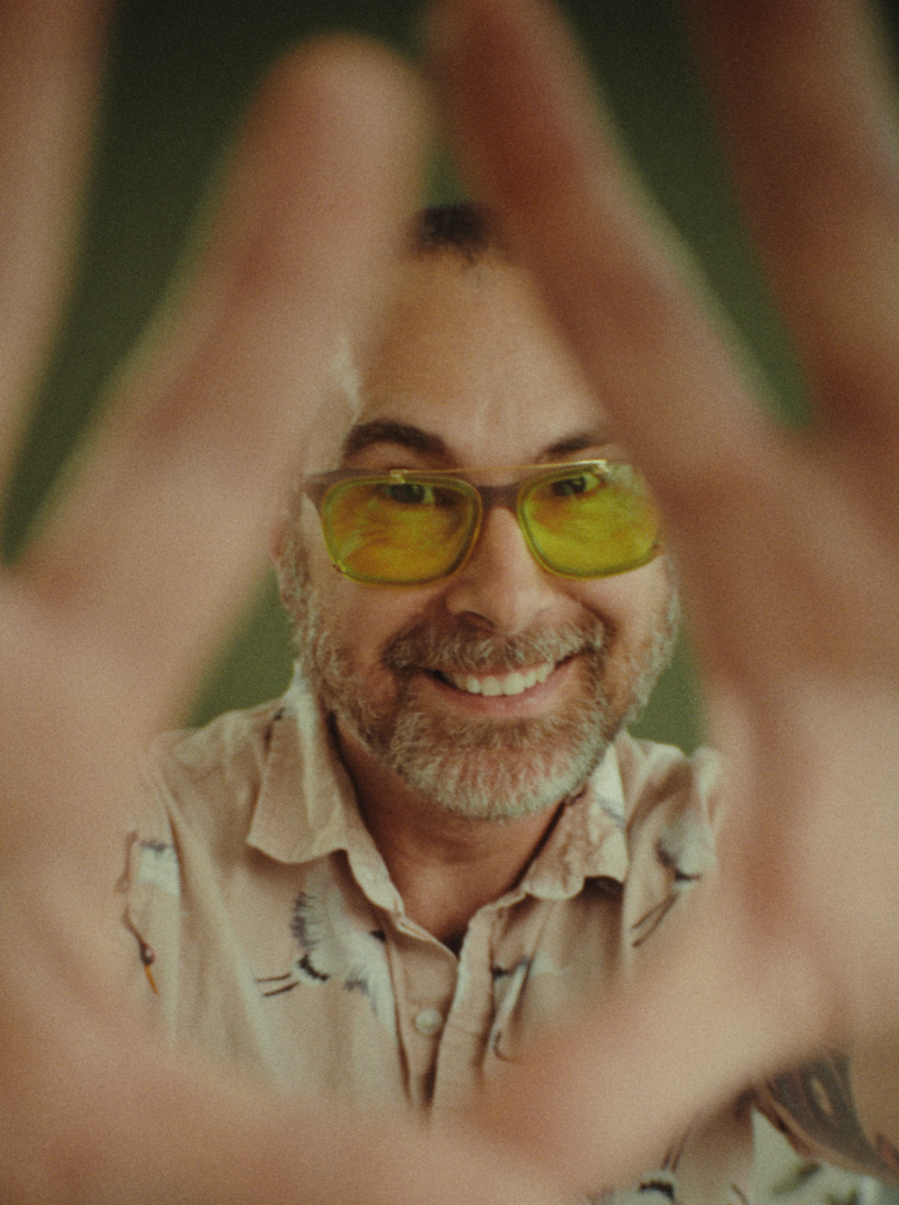 Domenic peeks through his hands. He's wearing yellow sunglasses, is smiling, has a short red/brown/grey beard and a light pink collared shirt with cranes.