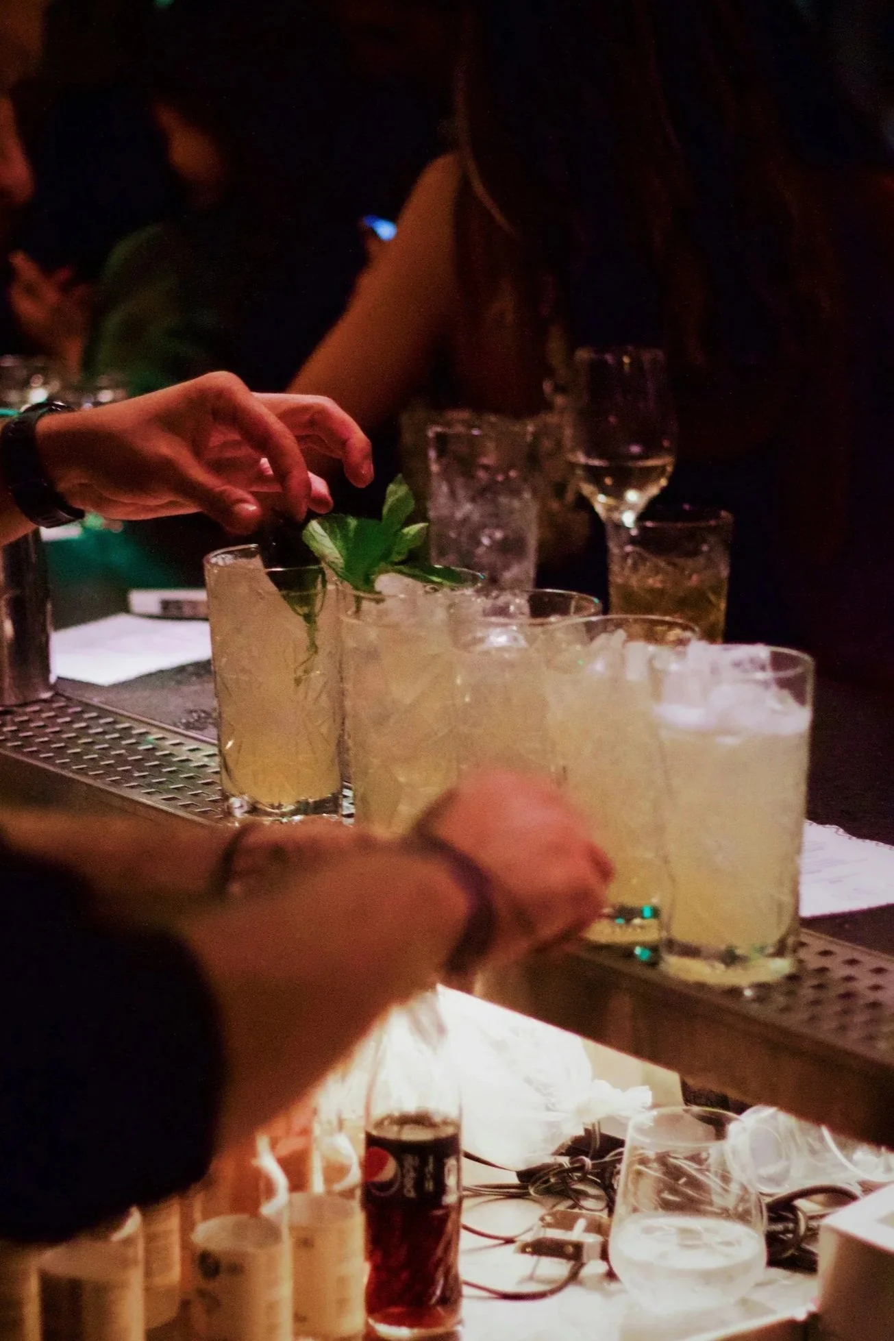 People preparing drinks at a bar or party, with glasses filled with beverages and garnishes on the counter.