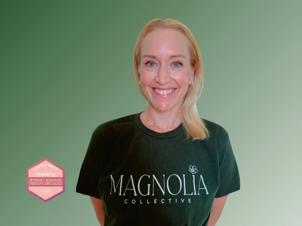 Portrait of a smiling woman with blonde hair, wearing a black top, pearl earrings, and a gold necklace with a small blue pendant. The background is green. There is a pink badge on her shoulder with the text 'Heather Hirsch Academy Women's Hormone Therapy Specialist'.