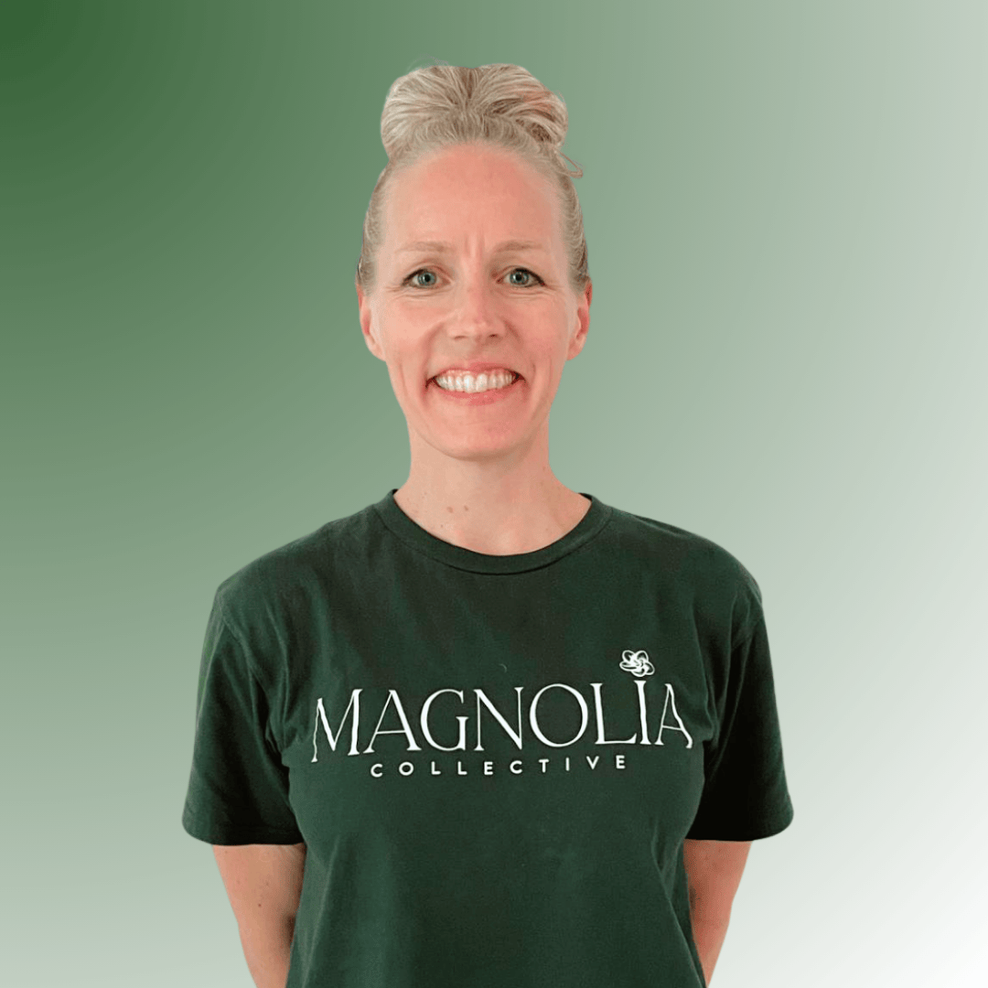 A smiling woman with curly blonde hair wearing a navy sleeveless top and a necklace with a small pendant, standing against a green background.