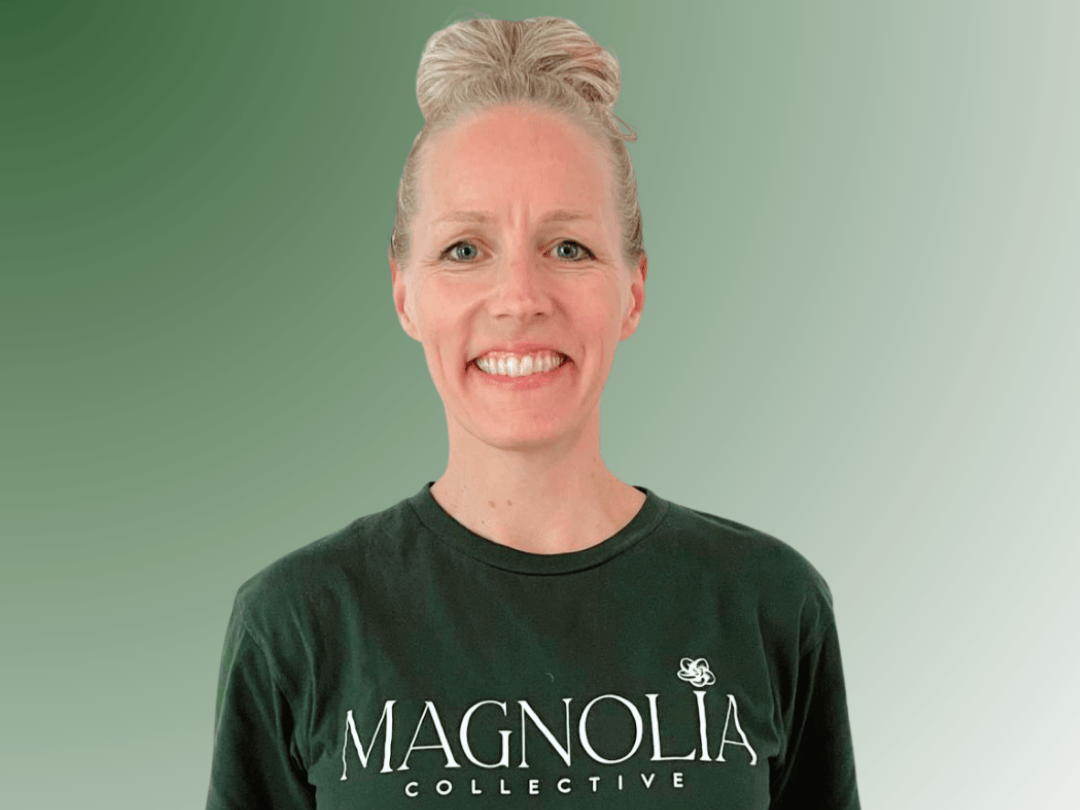 A smiling woman with curly blonde hair wearing a navy sleeveless top and a necklace with a small pendant, standing against a green background.