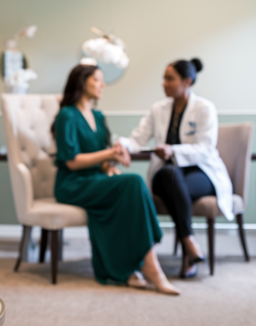 A woman in a green dress sitting on a beige chair, holding hands with a woman in a medical coat, sitting on a similar chair, in a consultation or therapy setting.