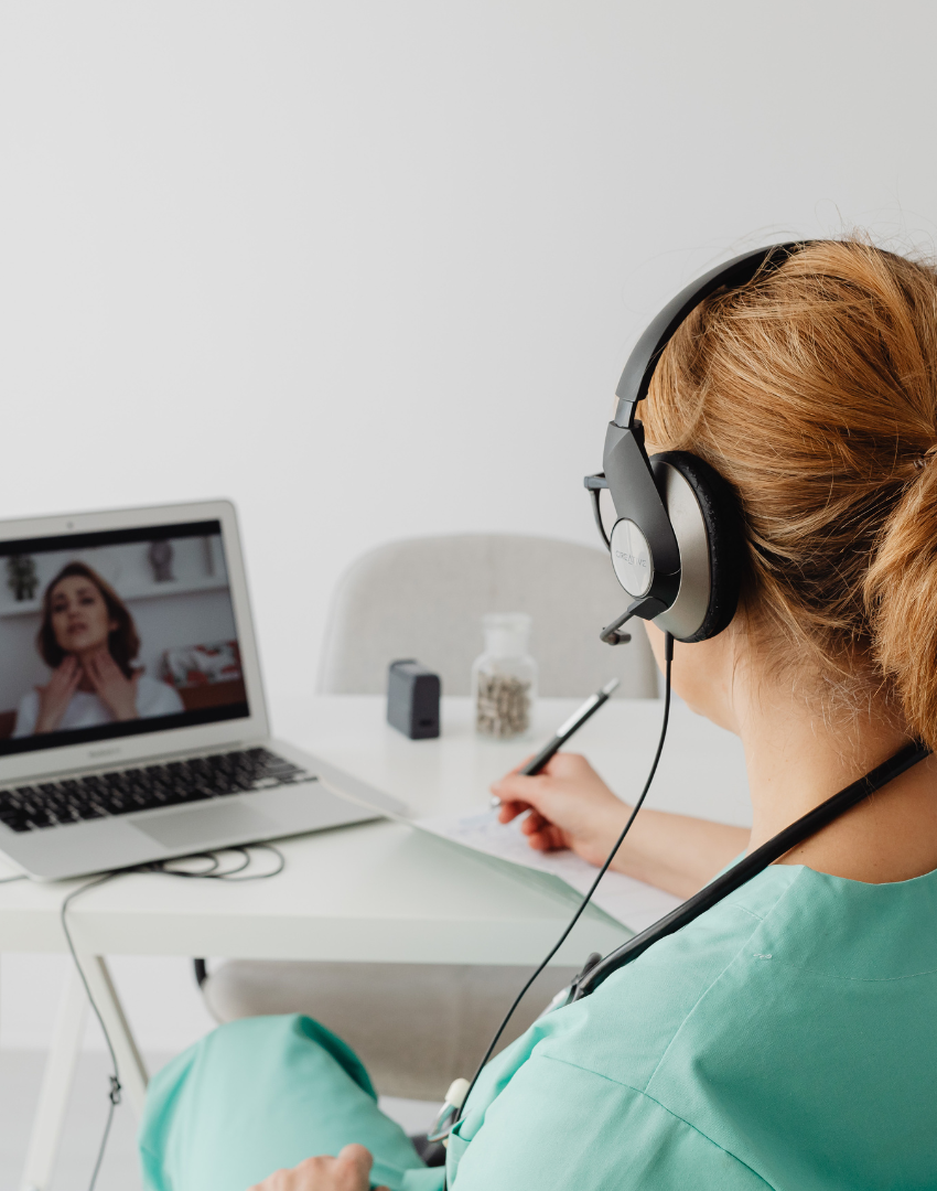 A nurse or healthcare worker with red hair wearing scrubs and a headset, on a virtual consultation with a woman on a laptop, in a clinical or home setting.