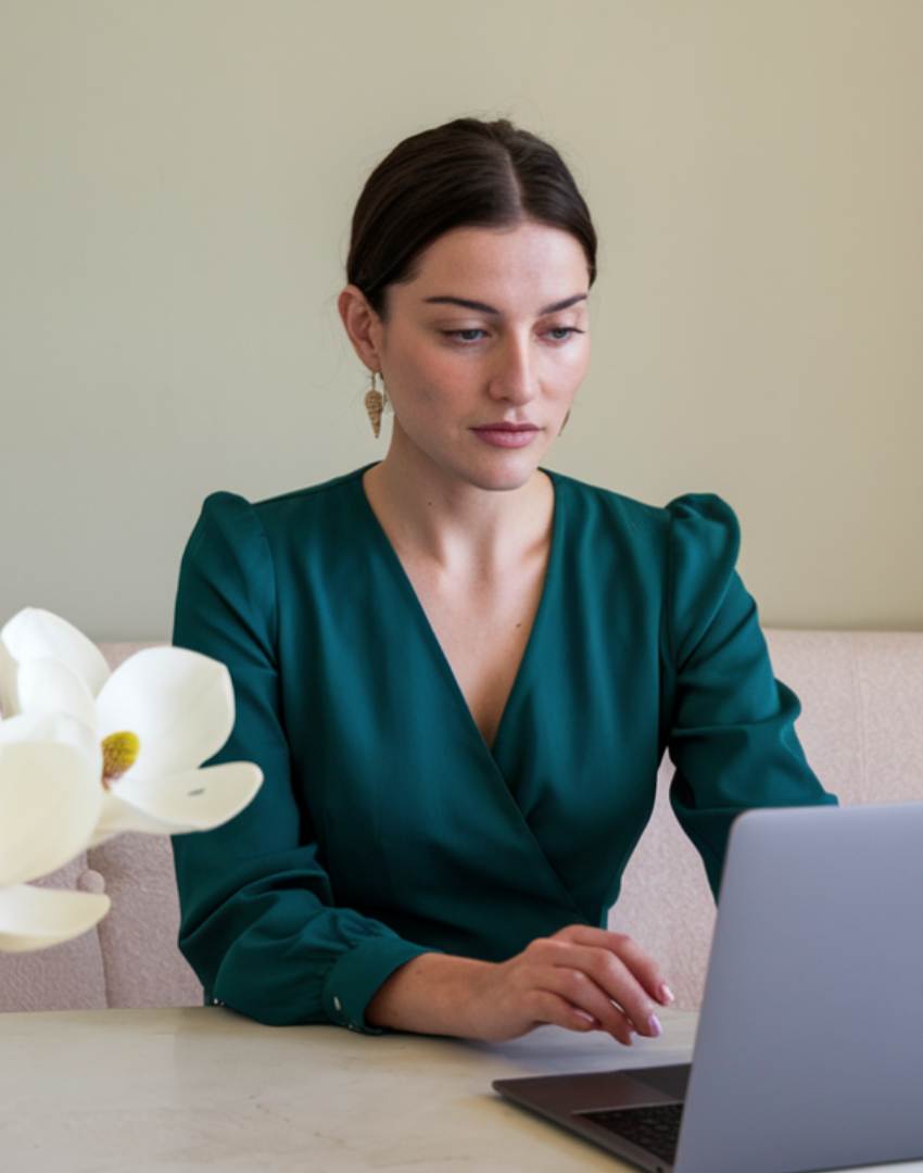 Woman with dark brown hair in a bun, wearing a teal blouse and earrings, working on a laptop at a table with a white flower in the foreground.