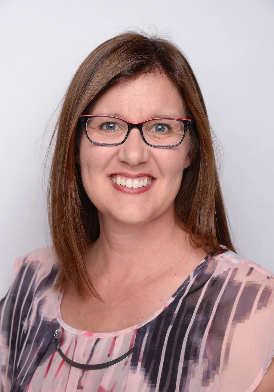 A woman with brown hair, wearing glasses and a patterned blouse, smiling at the camera against a plain light background.