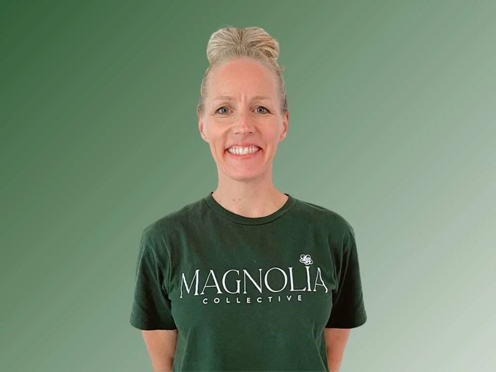 A smiling woman with curly blonde hair wearing a navy sleeveless top and a necklace with a small pendant, standing against a green background.