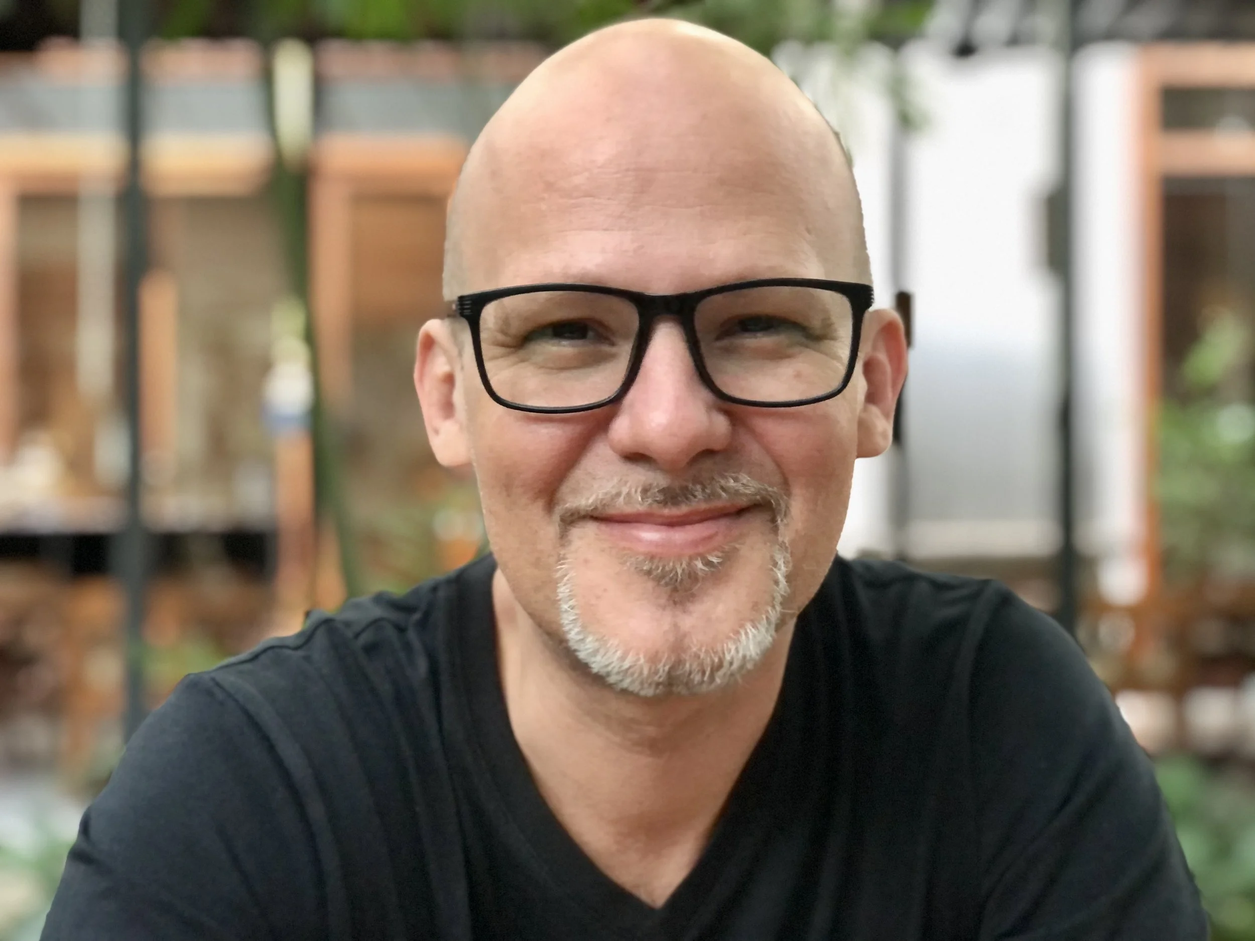 A man with a shaved head, black glasses, and a goatee smiling at the camera, sitting indoors with shelves and plants in the background.