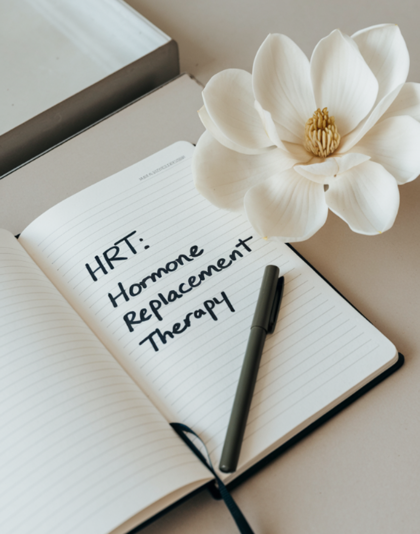 Open notebook with handwritten text reading 'HRT: Hormone Replacement Therapy', a black pen resting on the page, and a white magnolia flower beside it on a light-colored surface.
