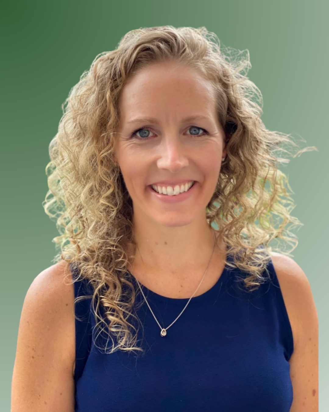 A smiling woman with curly blonde hair wearing a navy sleeveless top and a necklace with a small pendant, standing against a green background.