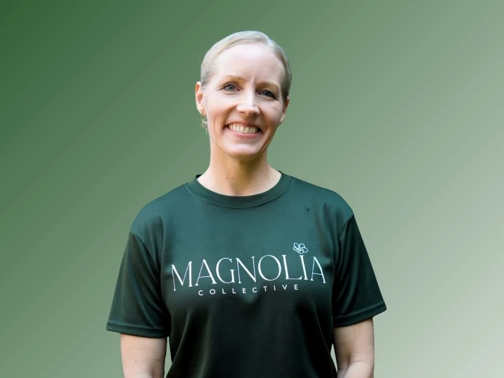 A smiling woman with curly blonde hair wearing a navy sleeveless top and a necklace with a small pendant, standing against a green background.