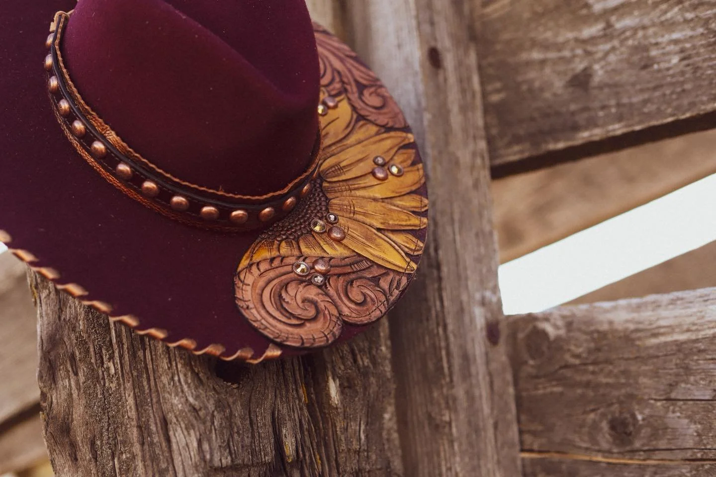 A maroon western hat with decorative studded band and a wide, sculpted leather brim, resting on a weathered wooden surface.