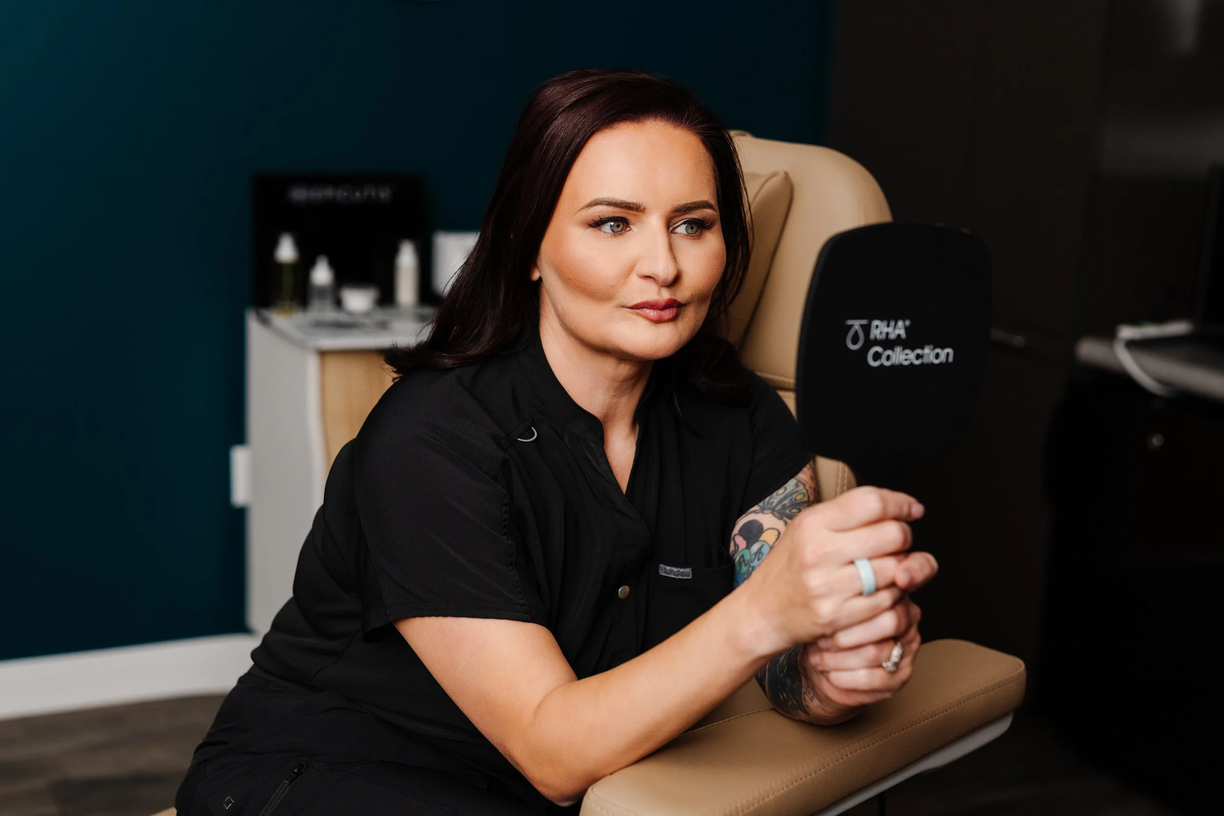 A woman with dark brown hair and tattoos on her arm is sitting on a beige chair, looking at a black screen with the words 'RHA Collection' on it, in a professional setting.