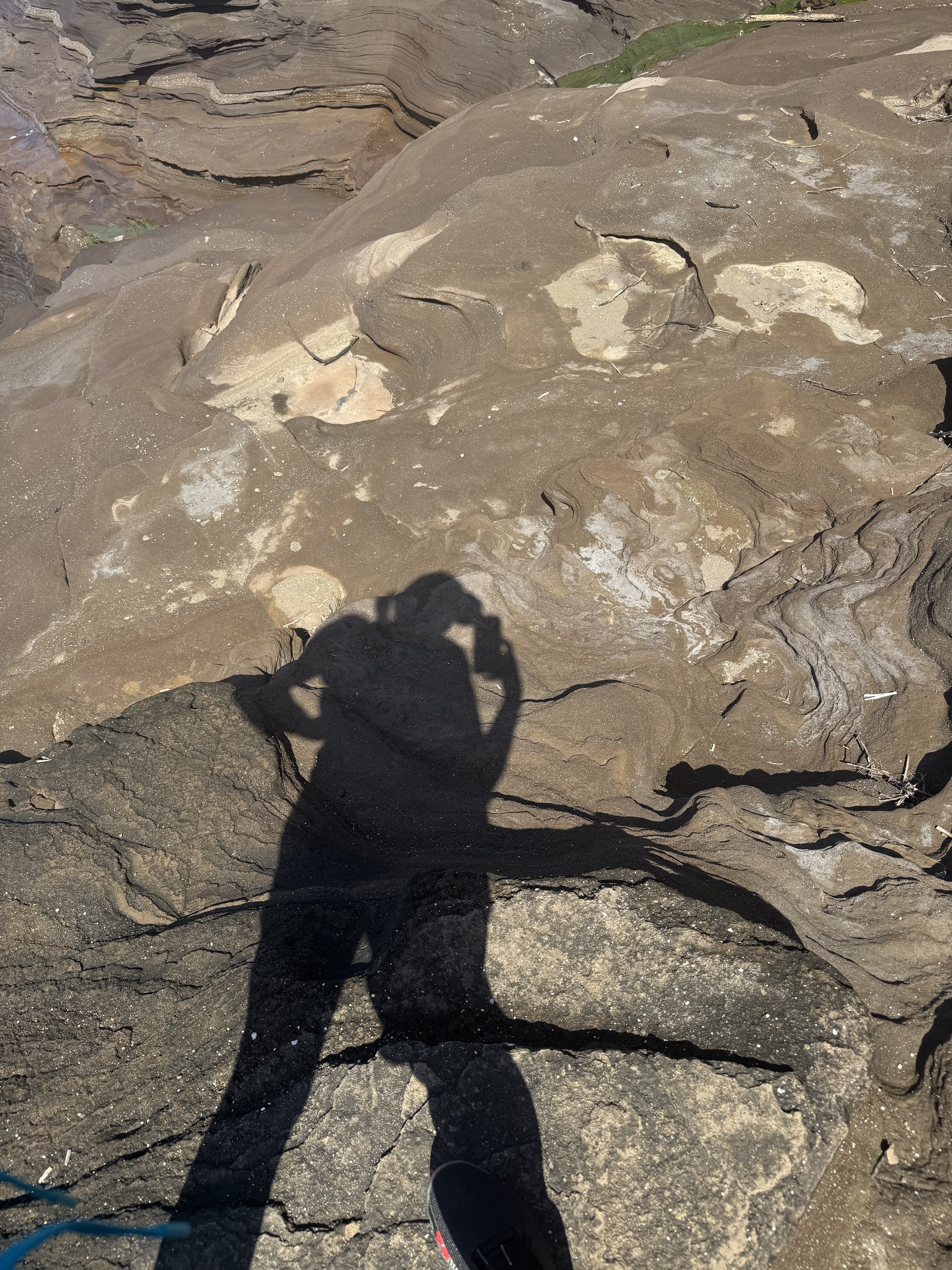 A person standing on a rocky, uneven, sedimentary rock surface with visible layers and cracks, their shadow cast on the ground. Part of a shoe is visible at the bottom of the image.