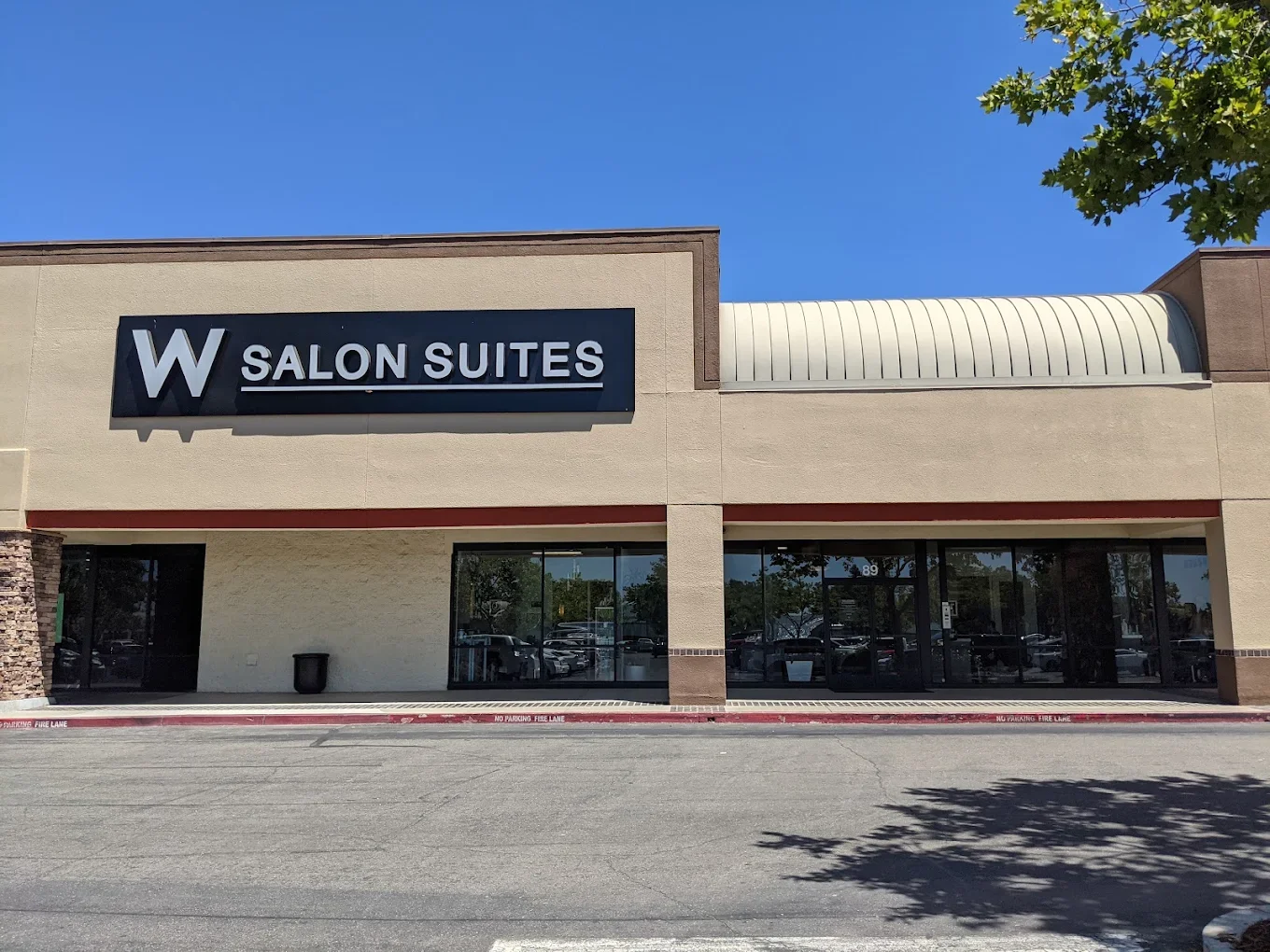 Exterior view of a strip mall with a sign reading 'Salon Suites' under a large letter 'W', a row of windows, and a shaded parking lot in front.