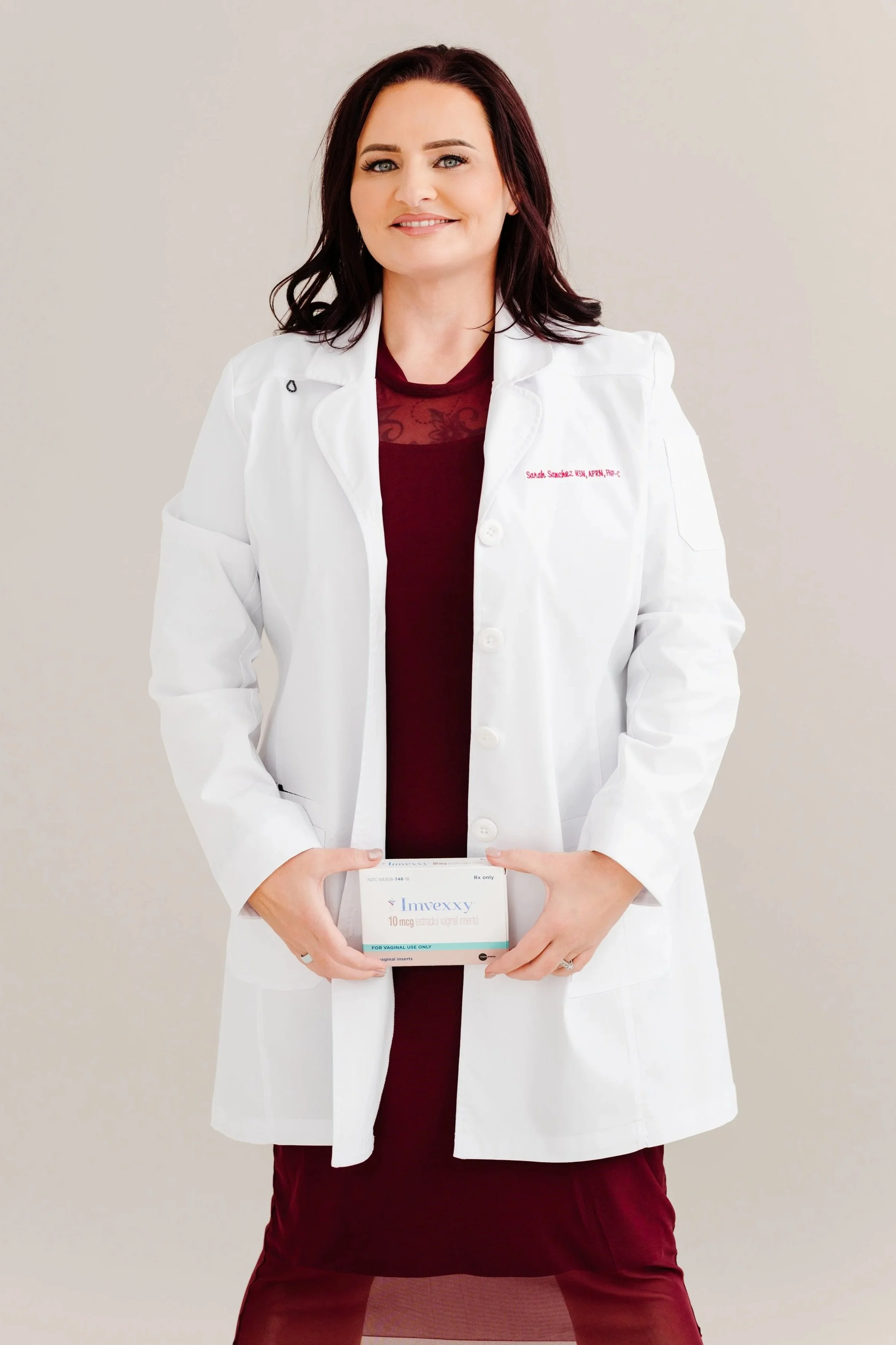 A woman in a white doctor’s coat holding a box of Invexxy vaginal inserts, standing against a plain light-colored background.