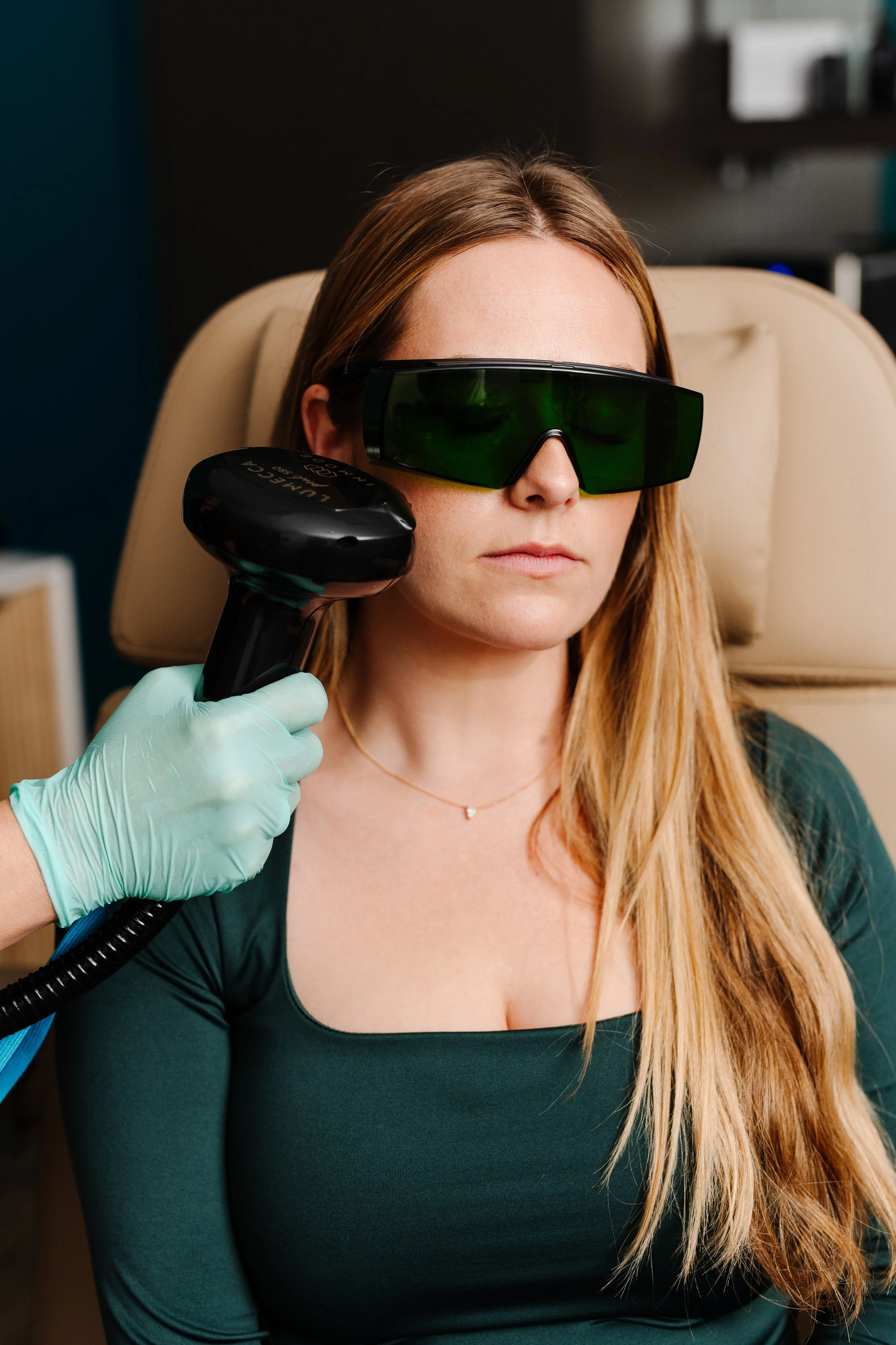 A woman with long blonde hair sitting in a medical chair, wearing green protective sunglasses. A person wearing a green glove is holding a laser device close to her face, possibly undergoing a cosmetic or dermatological procedure.