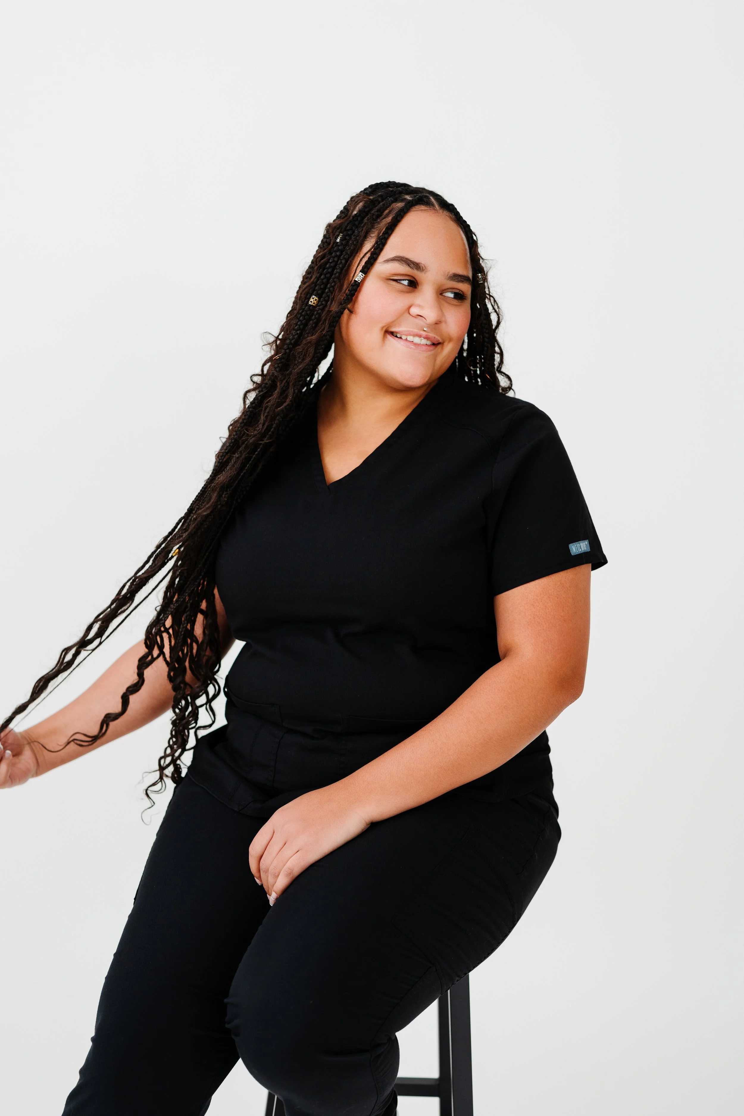 A woman with long braided hair, wearing black scrubs, sitting on a black stool against a plain white background, smiling and looking to the side.