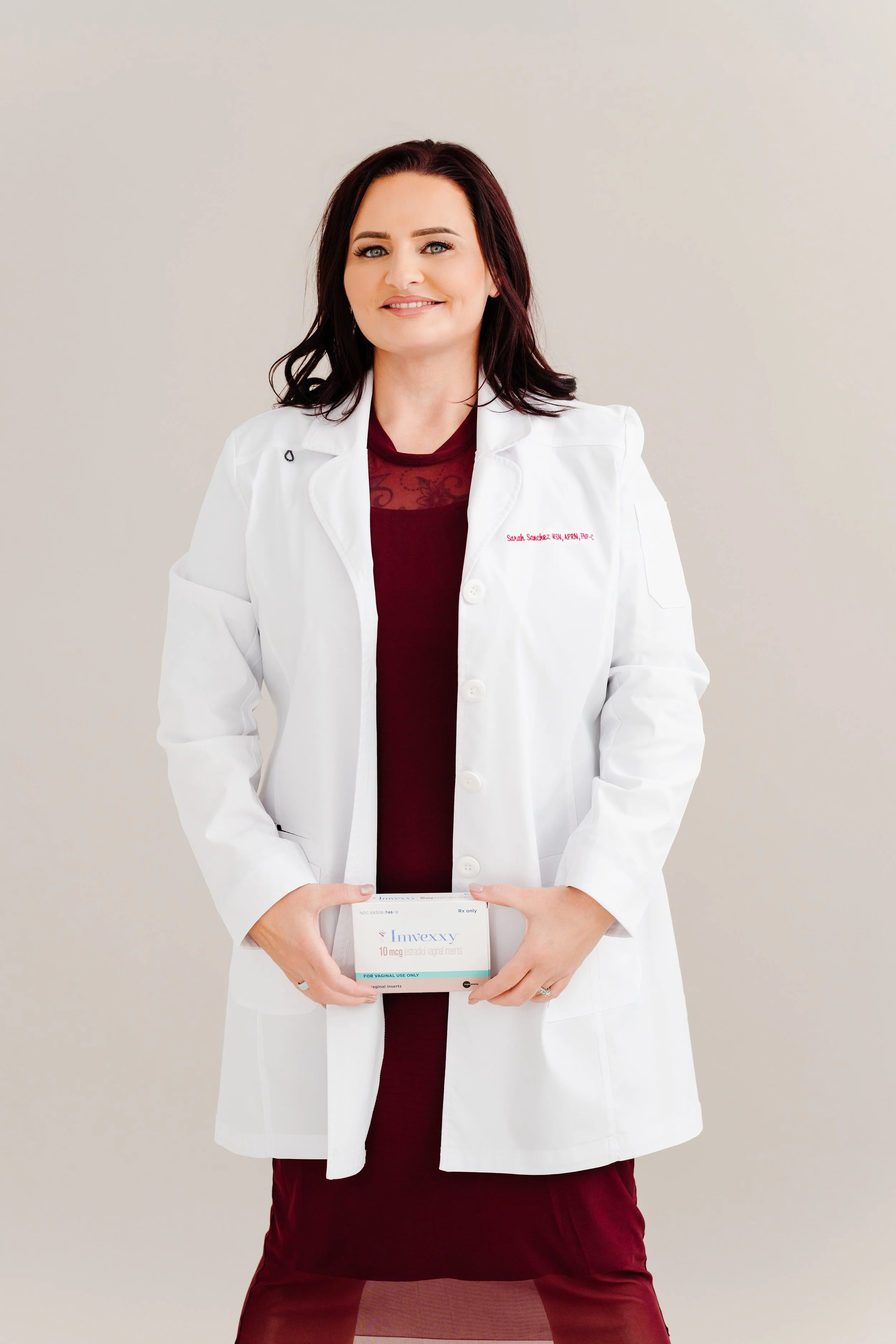 A woman in a white doctor's coat holding a box of medication, standing against a plain light-colored background. The coat has red embroidered text and she has shoulder-length dark hair.