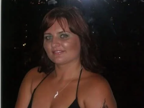 A woman with dark brown hair, wearing a black sleeveless top, standing outdoors at night with city lights in the background.