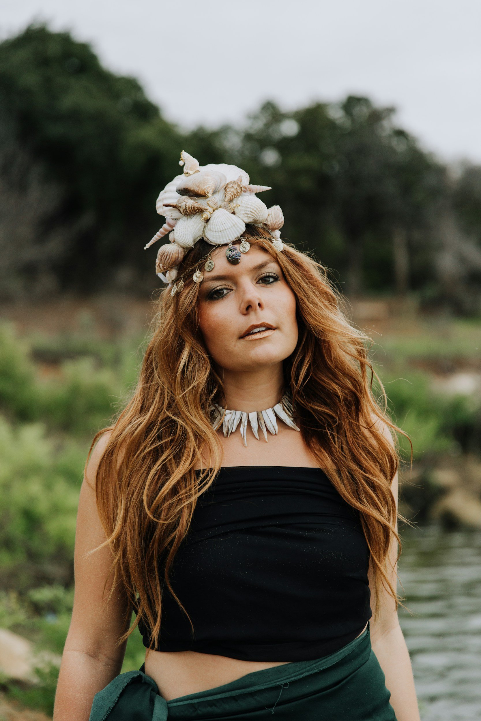 Woman in black top with seashell crown and necklace, standing outdoors.