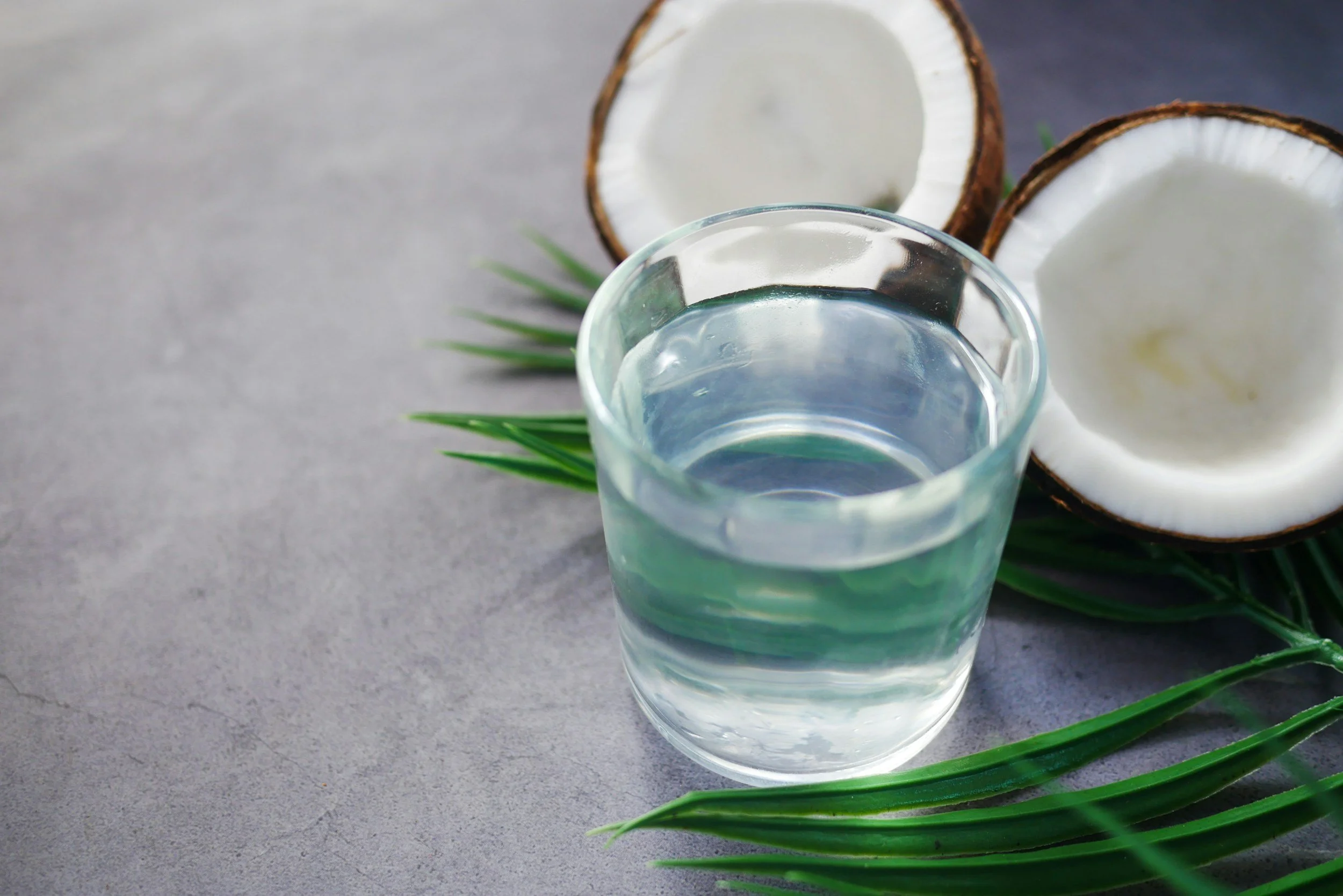 A clear glass of water is placed on a gray surface. Behind it, a halved coconut with white inner flesh and brown outer shell is visible, along with some green leaves and blades of grass.