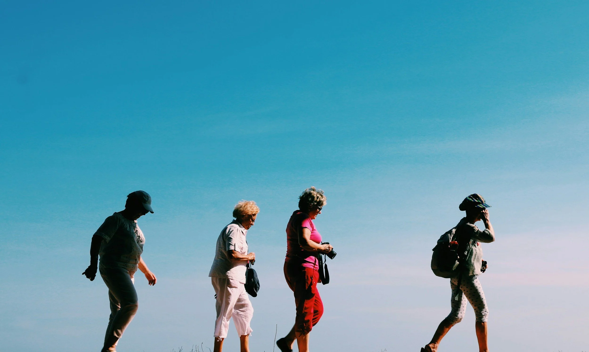 Group of five elderly women walking outdoors against a clear blue sky, some wearing hats and carrying cameras.
