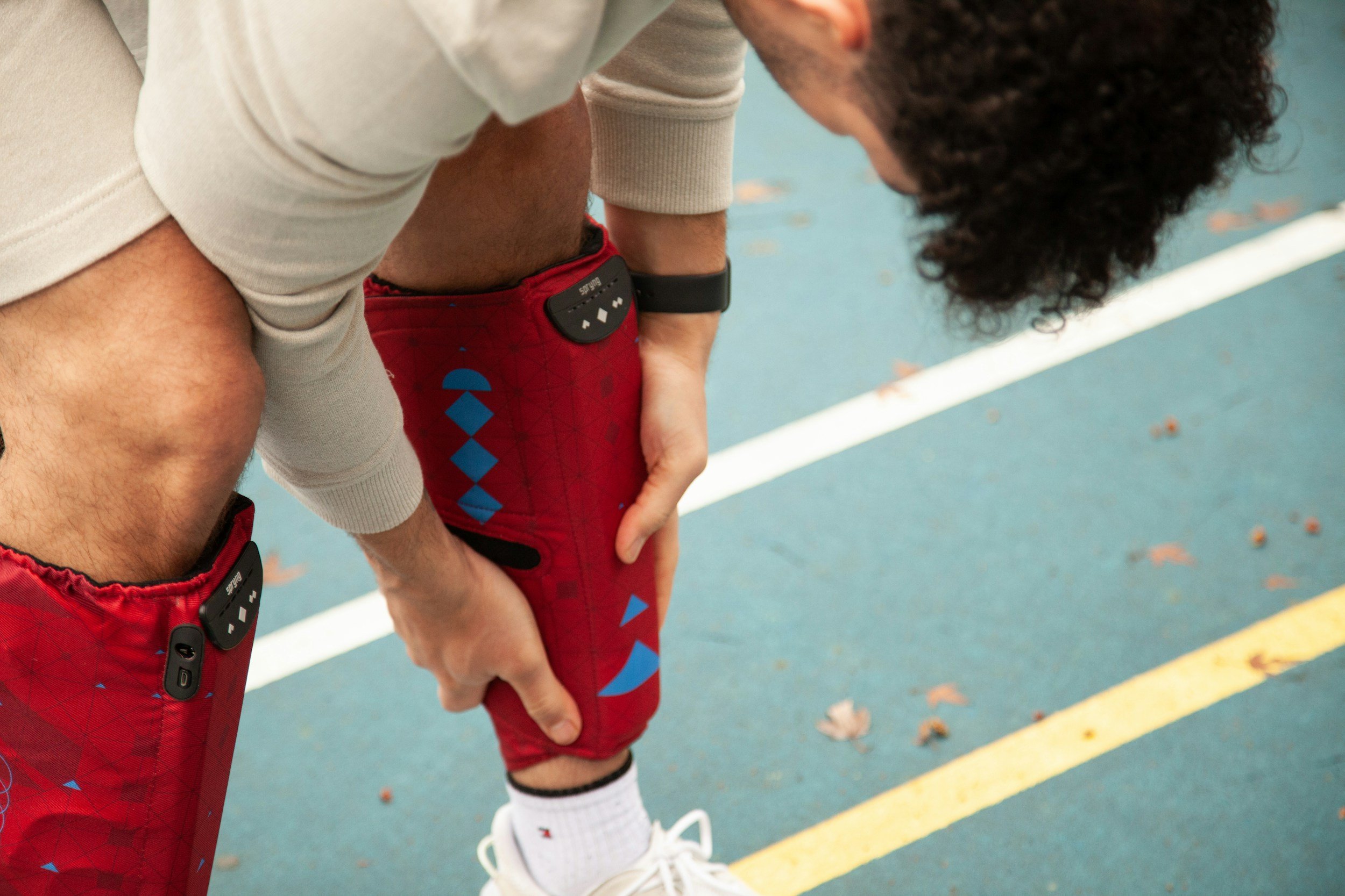 A person wearing a white short-sleeve shirt, red shorts with blue diamond patterns, white socks, and white sneakers, is bending over in athletic attire on a blue outdoor running track, adjusting their knee.