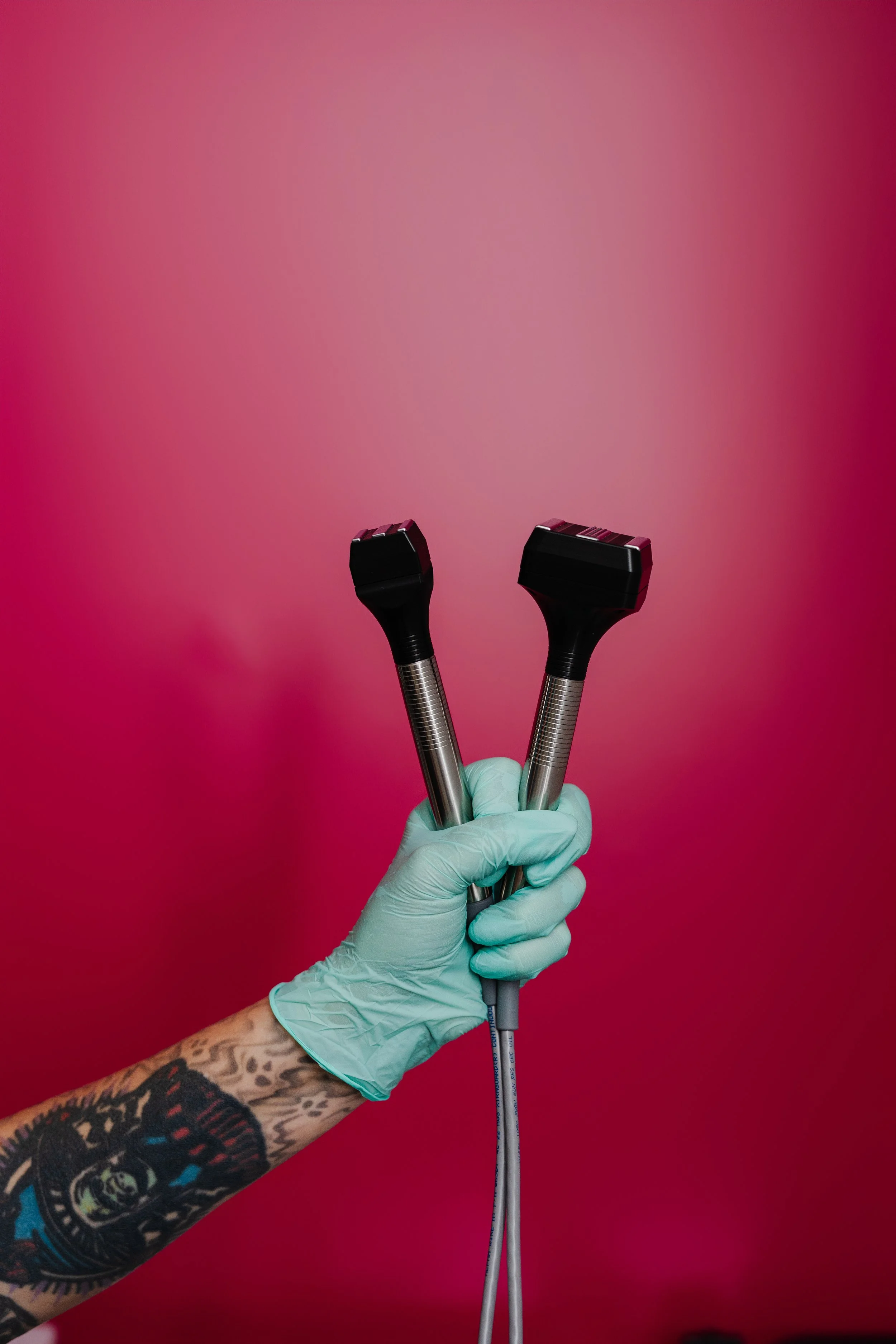A tattooed arm wearing a light green glove, holding two tattoo equipment needles, against a red background.