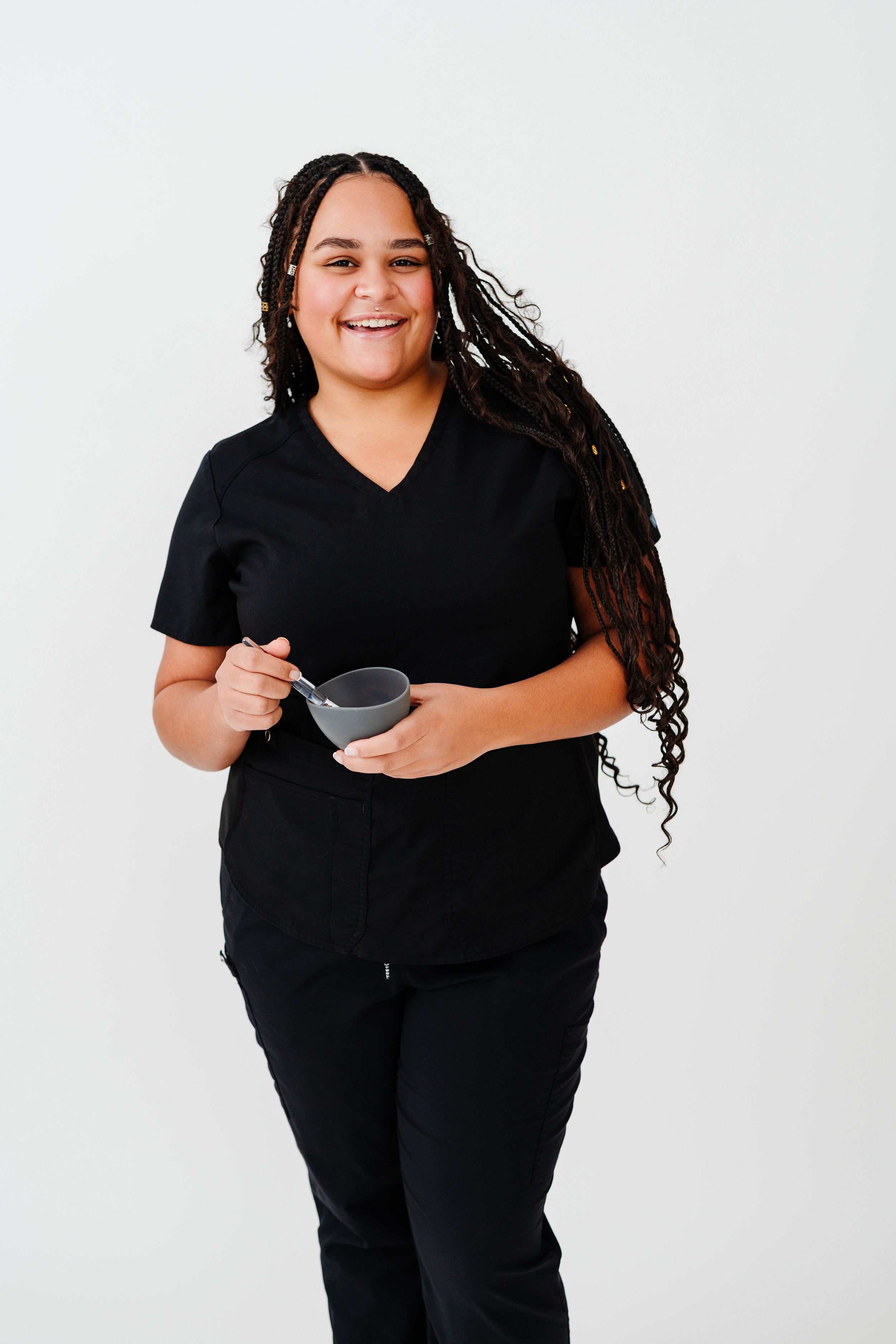 A smiling woman with long braided hair wearing black scrubs, holding a gray mug and a spoon, standing against a plain white background.