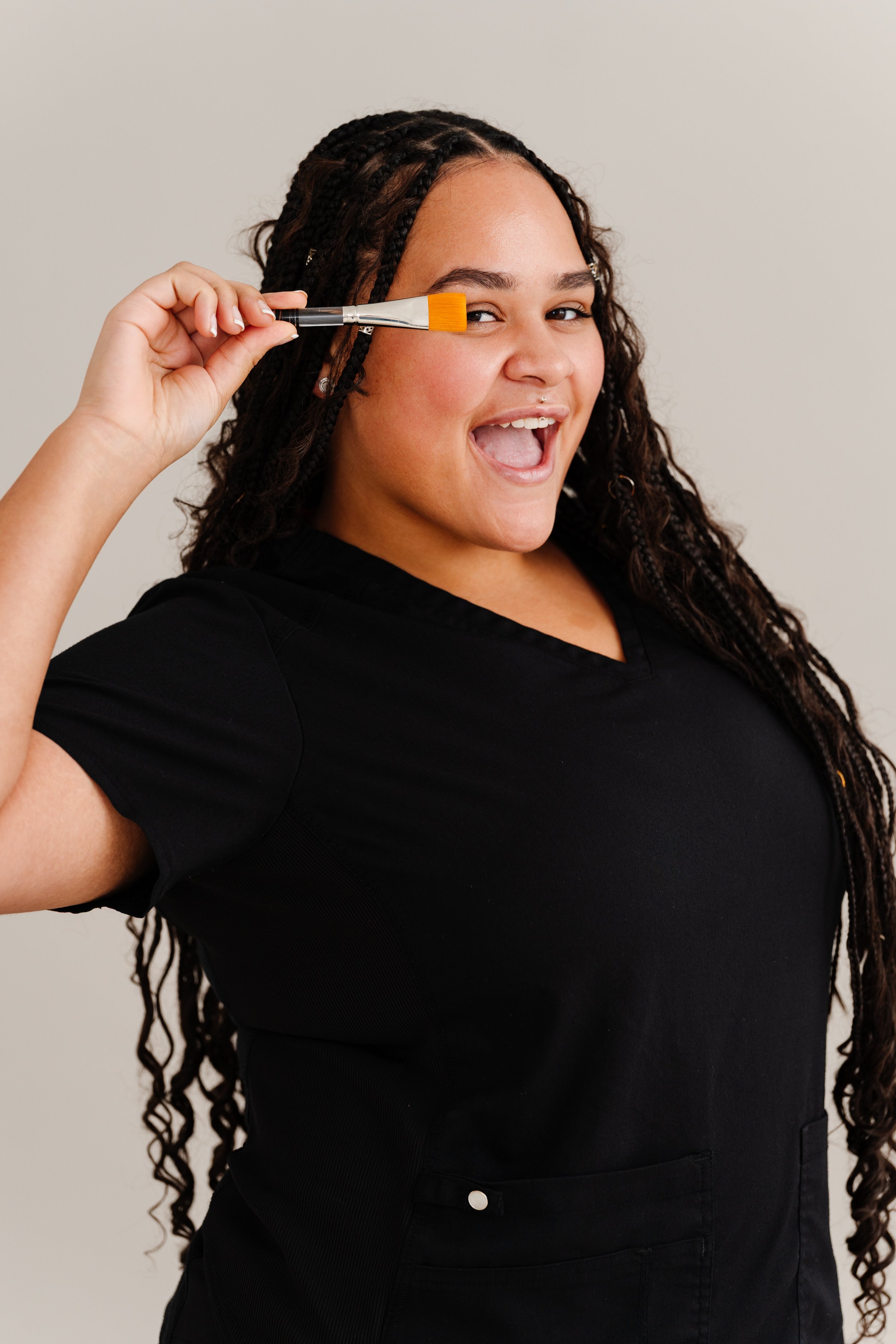 A woman with long, curly hair, smiling, holding a makeup brush near her face, wearing a black top, standing against a plain light-colored background.